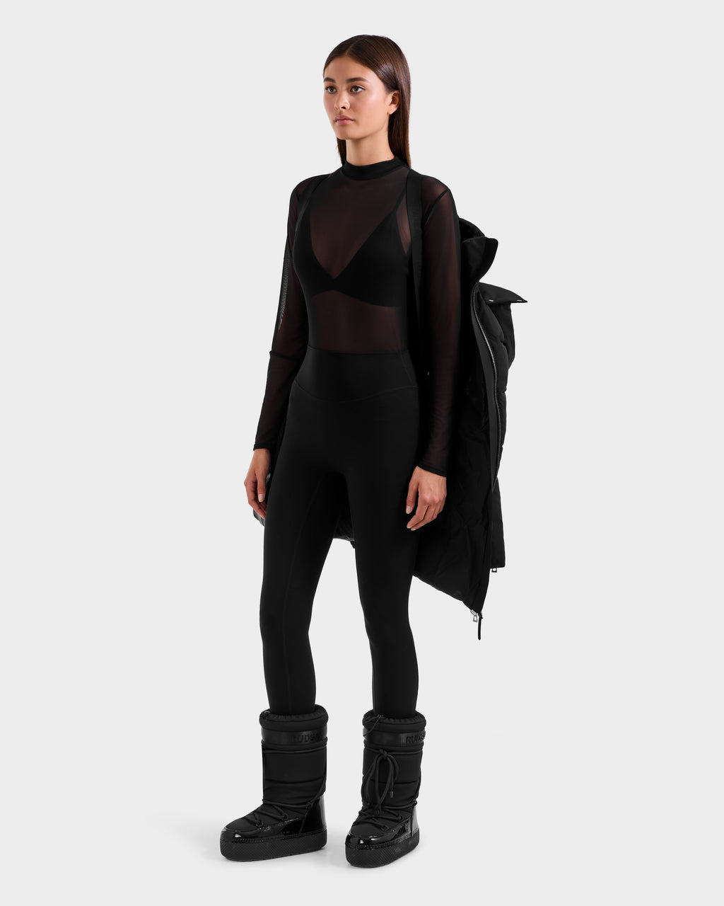 A woman poses against a plain background in the Rudsak CAT H - 8125588 Black down puffer draped over one shoulder, sheer black long-sleeve top over a bra, high-waisted black leggings, and black snow boots.