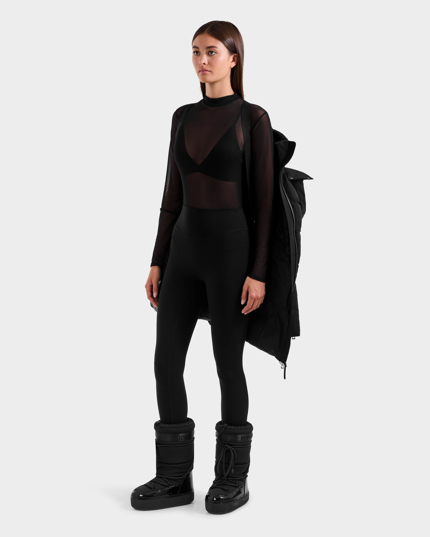 A woman poses against a plain background in the Rudsak CAT H - 8125588 Black down puffer draped over one shoulder, sheer black long-sleeve top over a bra, high-waisted black leggings, and black snow boots.