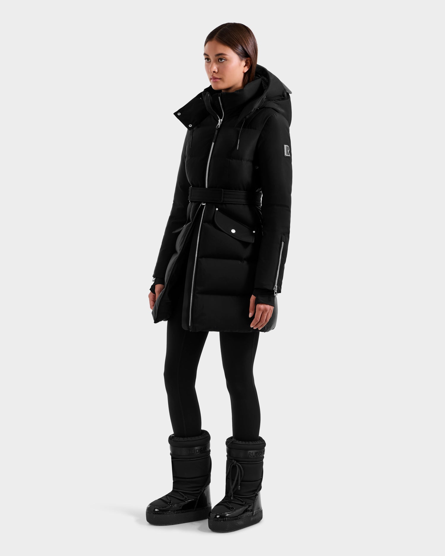 A woman wears the Rudsak CAT H - 8125588 Black RDS-certified down puffer coat with a hood, paired with black leggings and winter boots, standing against a light gray background facing slightly to the side with a neutral expression.