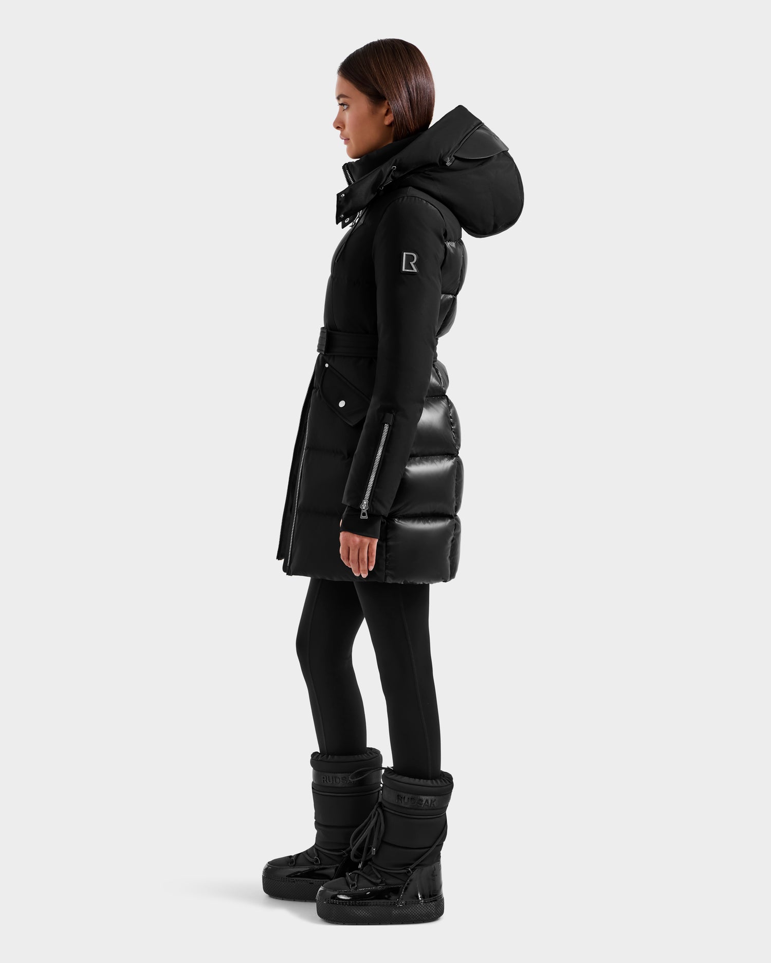A woman in the Rudsak CAT H - 8125588 Black RDS-certified down puffer coat with a hood, black leggings, and black winter boots stands in profile against a plain light gray background.