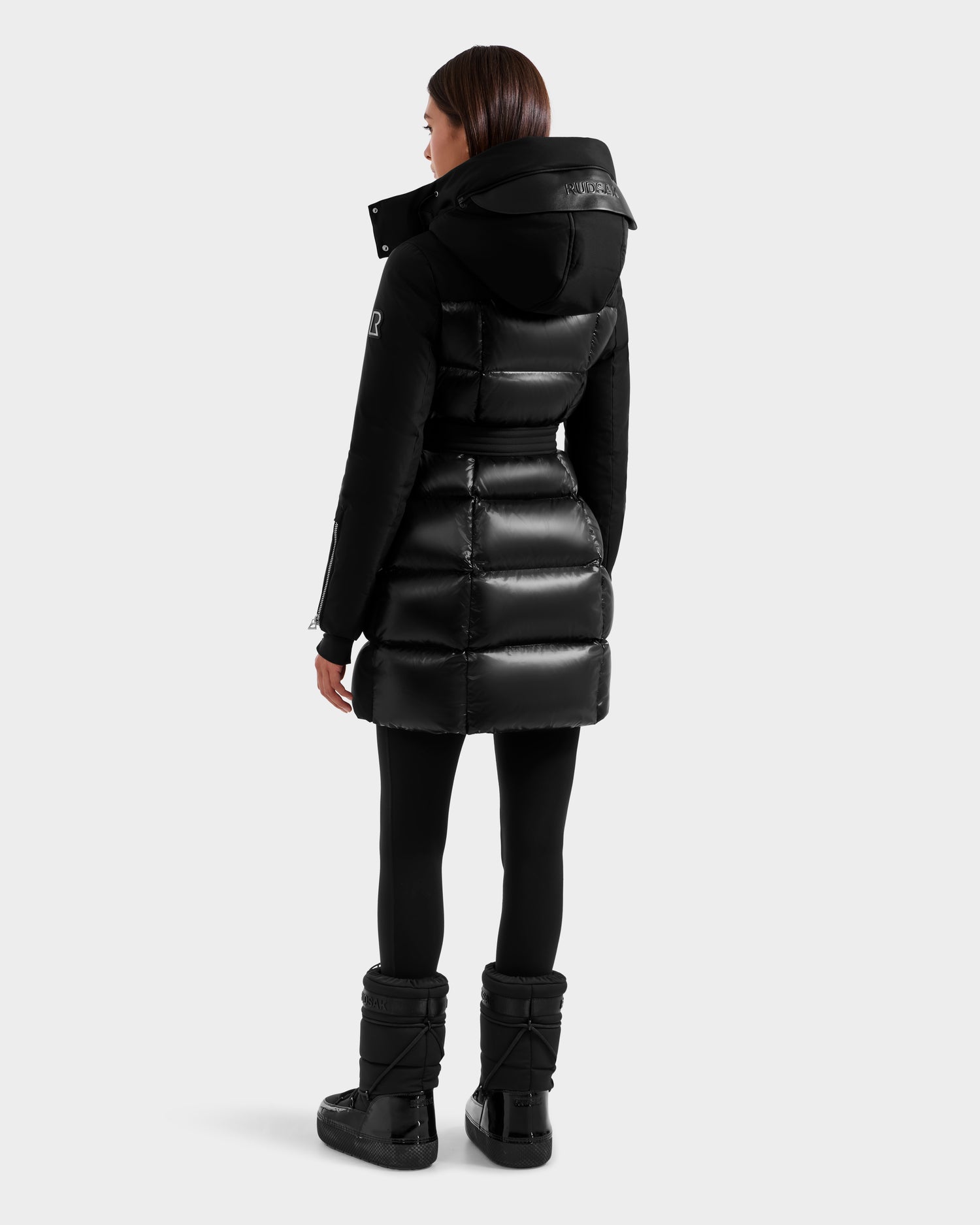 A woman faces away, wearing the Rudsak CAT H - 8125588 Black women's down puffer coat with hood, paired with black leggings and black winter boots, set against a plain light gray background.