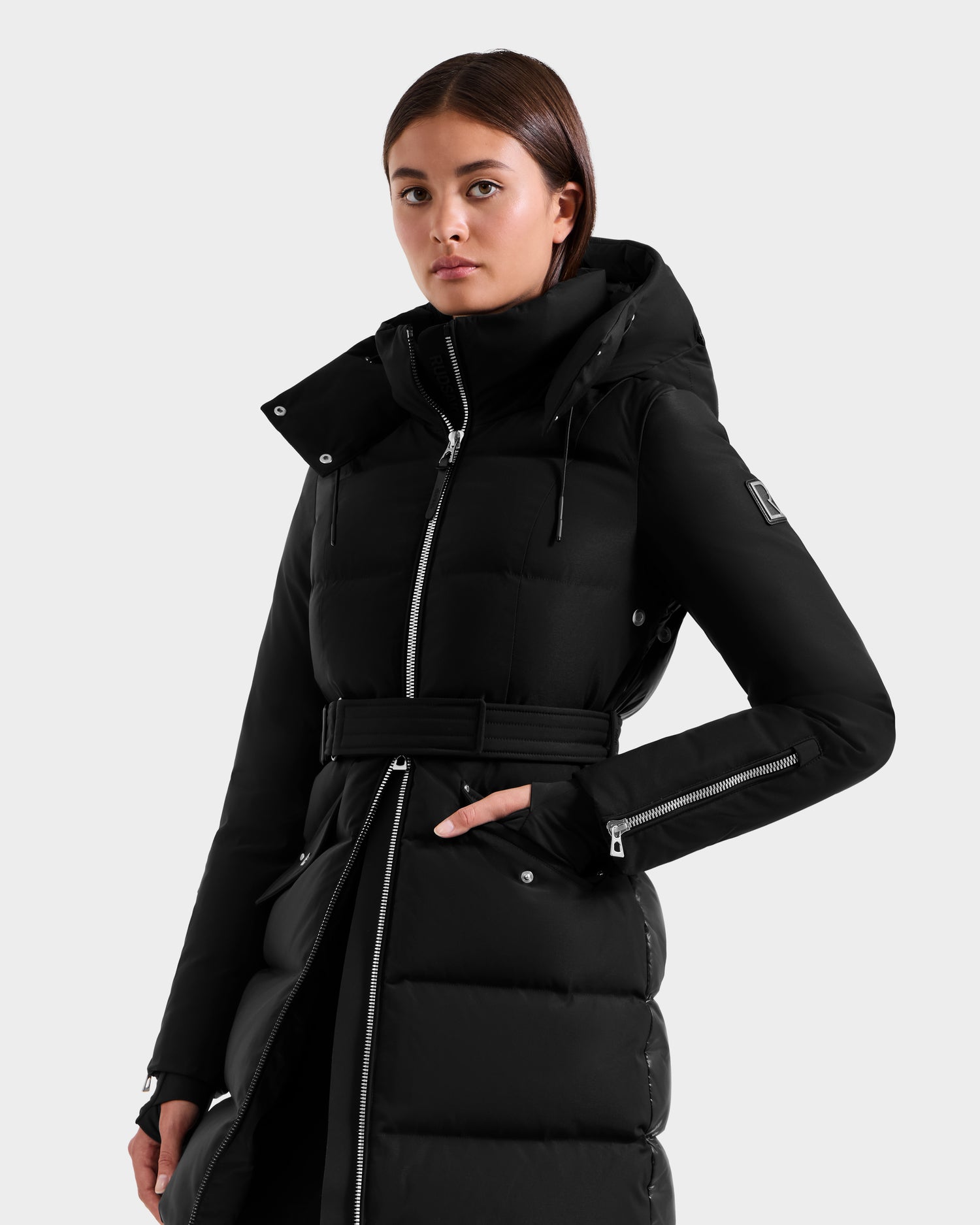 A woman with straight brown hair and a neutral expression stands against a light gray background, wearing the Rudsak CAT H - 8125588 Black fitted down puffer coat with hood—an effortless winter essential.