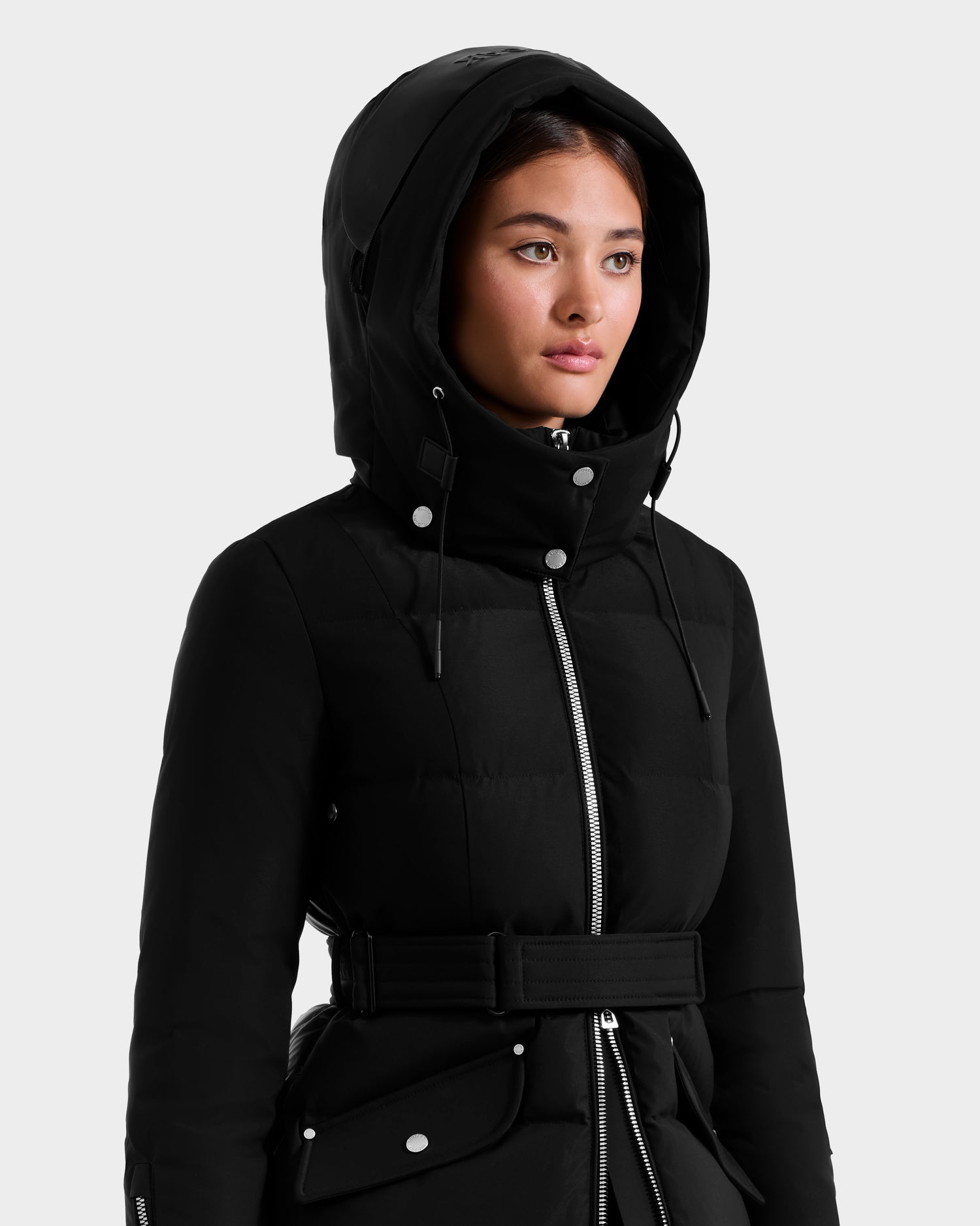 A woman wearing the Rudsak CAT H - 8125588 Black puffer jacket with a belt, silver zipper, and buttons stands against a plain light gray background.