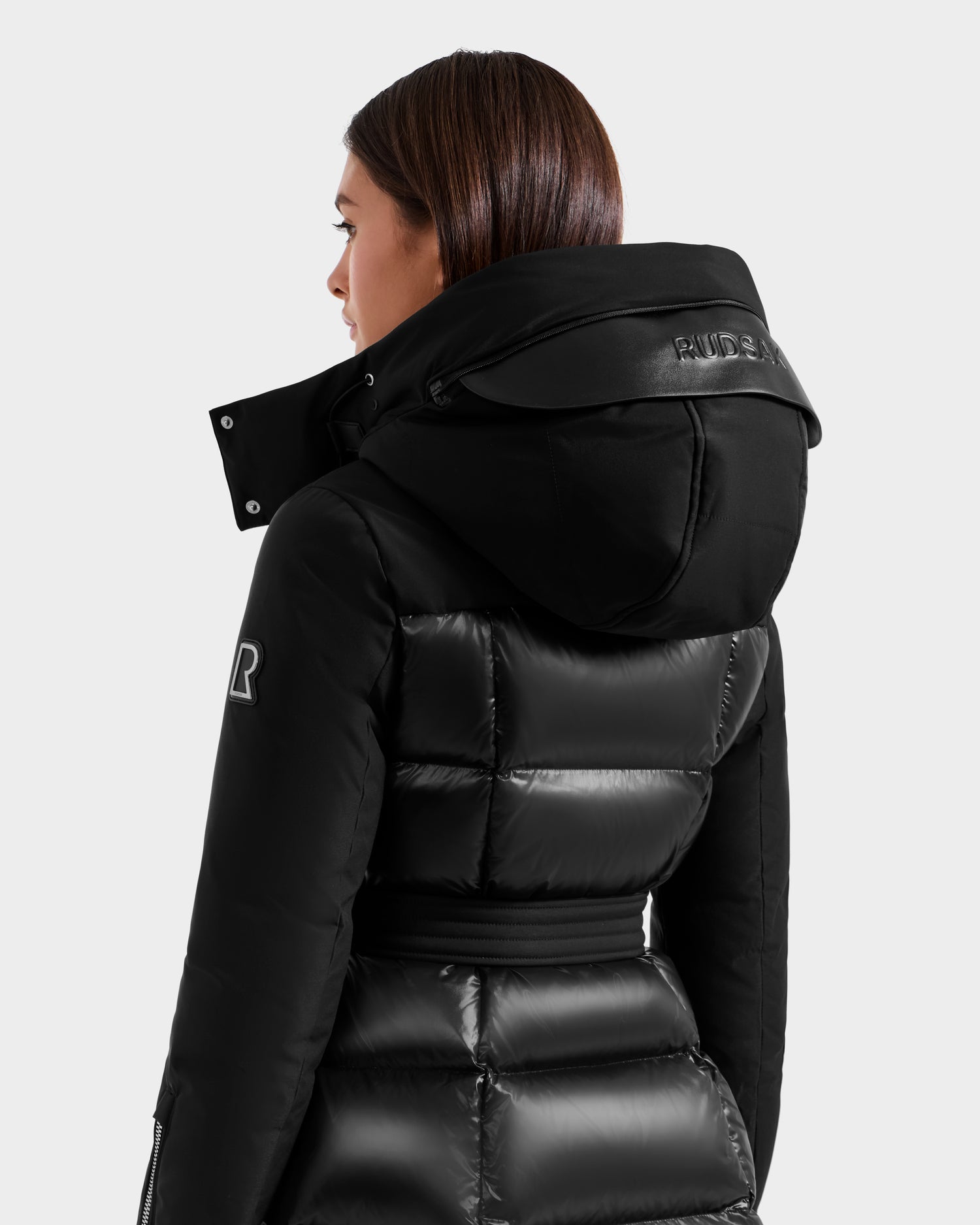 A woman with straight brown hair wears the Rudsak CAT H - 8125588 Black shiny women’s down puffer winter coat, featuring a large hood and Rudsak logos. Filled with RDS-certified down, she is shown from the back and side.