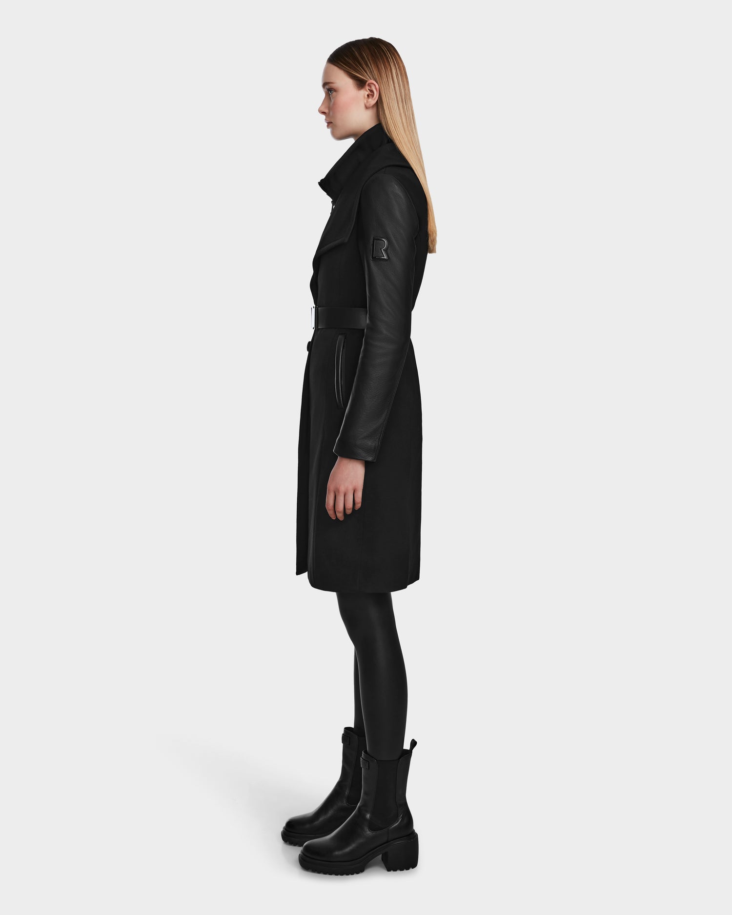 A woman with straight blonde hair stands in profile, modeling the Rudsak CELESTE - L124542 Black transitional coat with leather sleeves, paired with black leggings and chunky black boots against a plain white background.