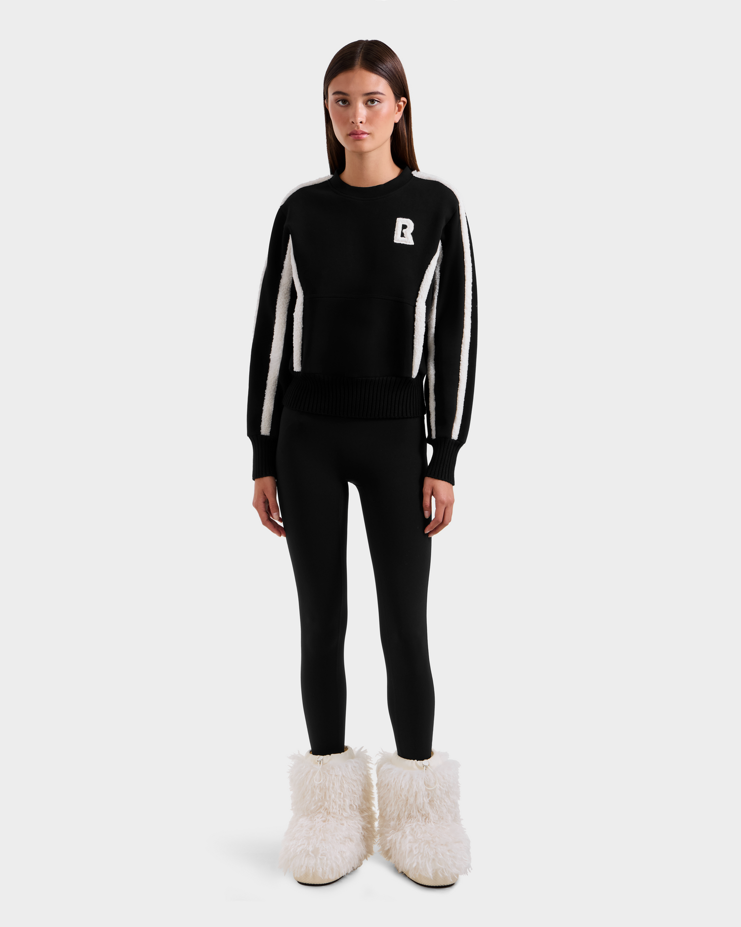 A woman poses against a plain background wearing the Rudsak CHANA - 8525656 Black women’s crew neck sweatshirt with a B logo, paired with black leggings and white furry boots for an après-ski chic look.