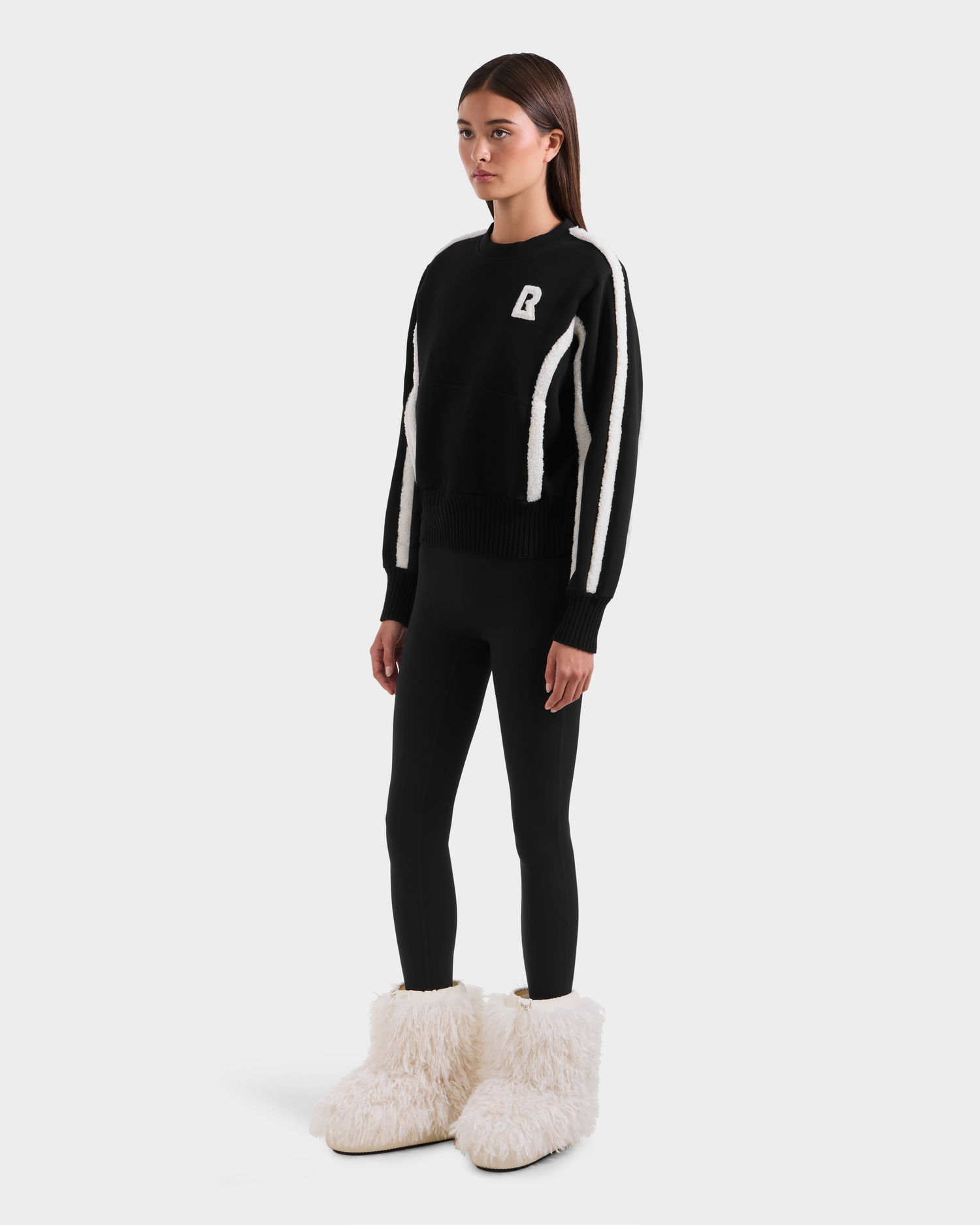A woman stands facing forward, wearing the Rudsak CHANA - 8525656 Black sweater with white stripes and a letter B on the chest, black leggings, and oversized fluffy white boots with faux shearling trim, against a plain white background.