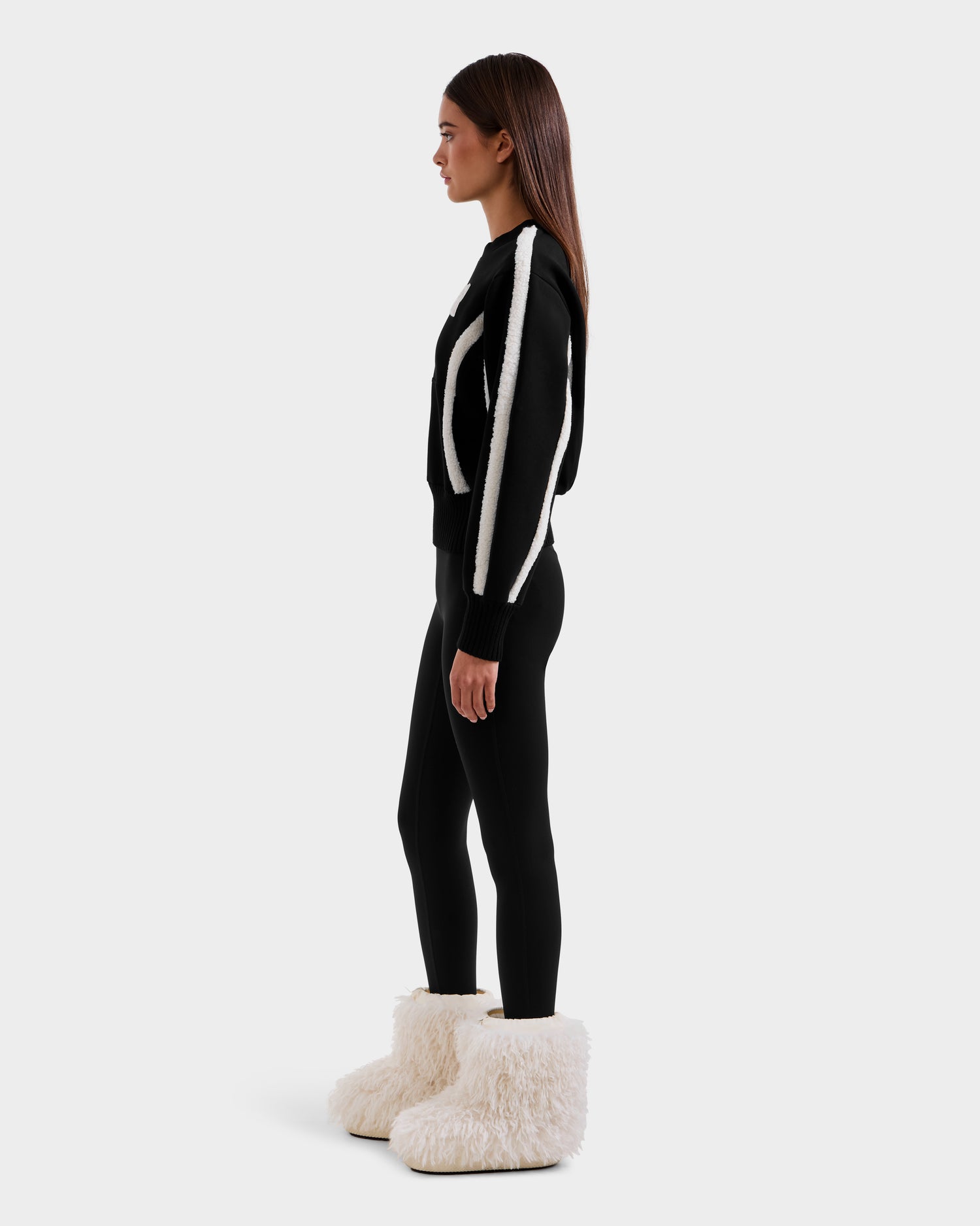A woman stands in profile wearing the Rudsak CHANA - 8525656 Black women’s crew neck sweatshirt with white stripes and large faux shearling-trimmed boots, creating an après-ski chic look against a plain white background.
