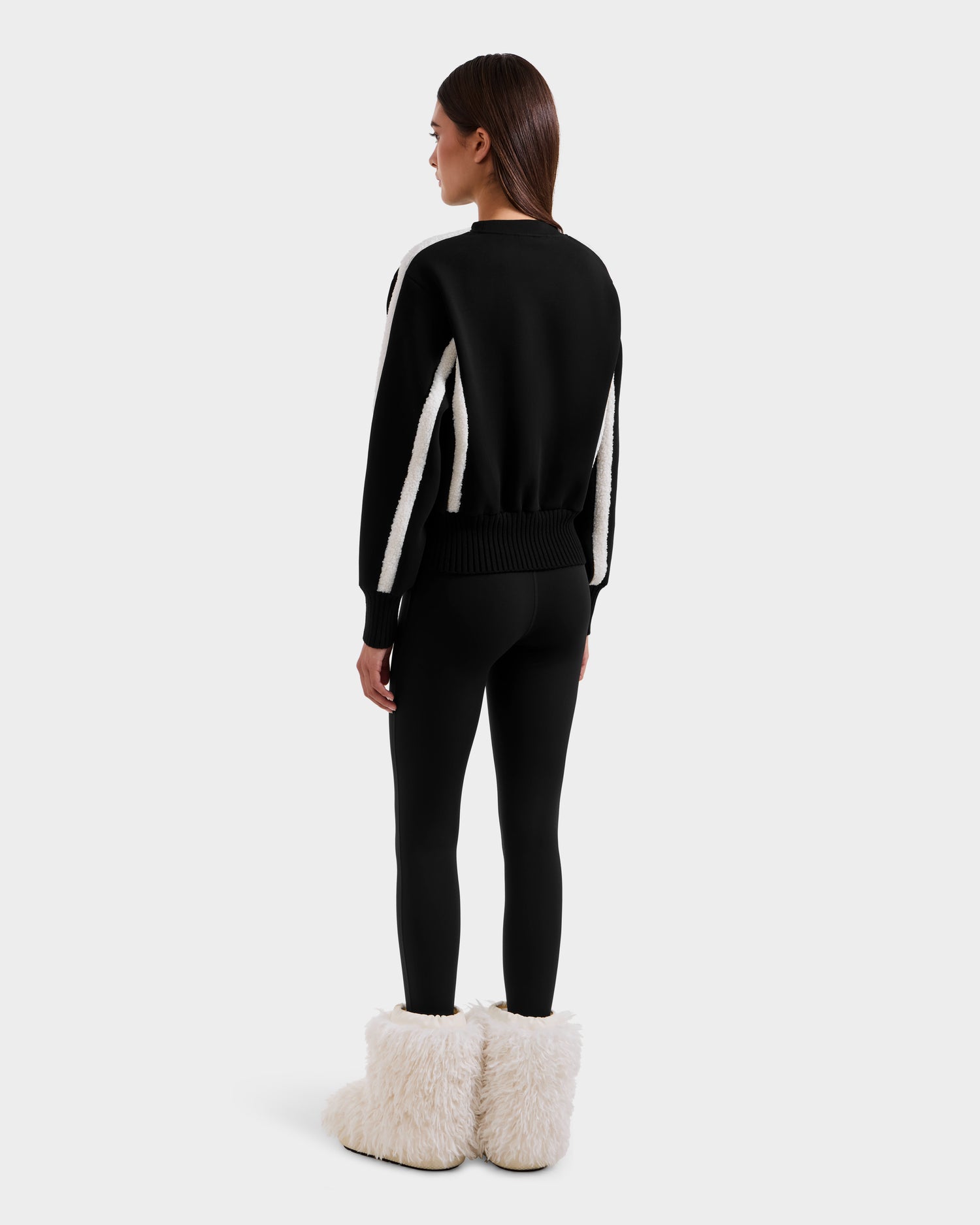 A woman faces away, wearing the Rudsak CHANA - 8525656 Black women’s crew neck sweatshirt with white sleeve stripes, black leggings, and white furry boots—showcasing a chic après-ski style against a simple light background.