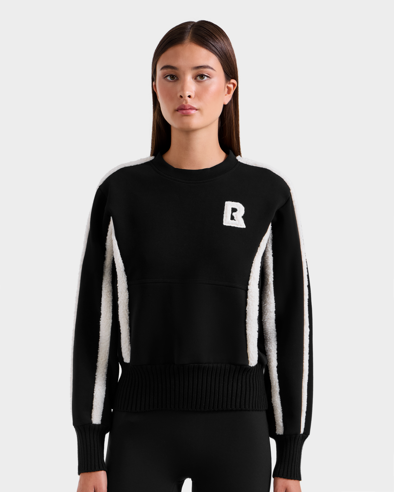 A young woman with straight brown hair models the Rudsak CHANA - 8525656 Black women's crew neck sweatshirt, featuring white textured stripes, faux shearling trim, and a white R logo on the chest, against a light gray background.