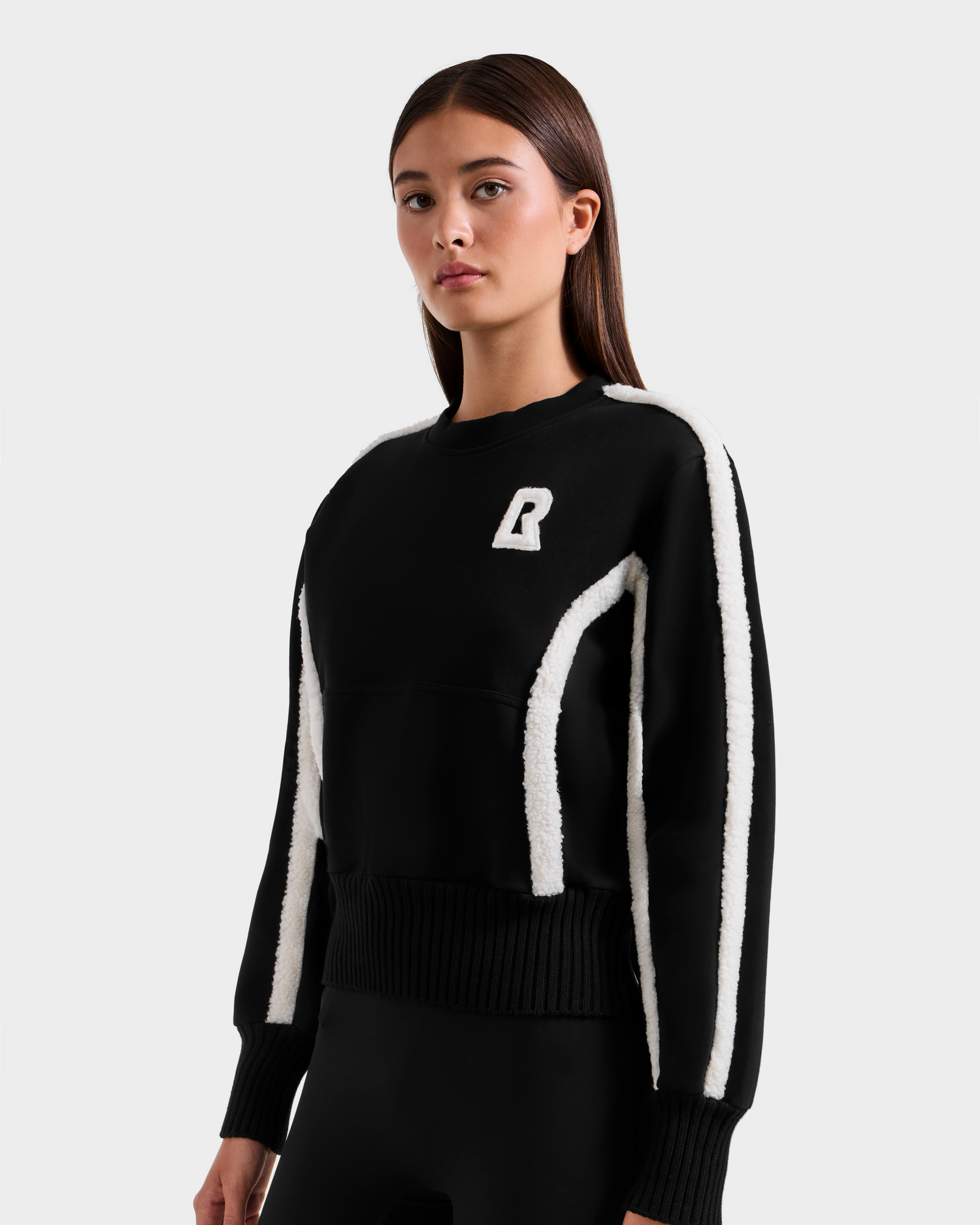 A woman stands against a plain light background, wearing the Rudsak CHANA - 8525656 Black women’s crew neck sweatshirt with white stripes on the sleeves and sides, and a bold white R emblem on the chest for après-ski chic style.