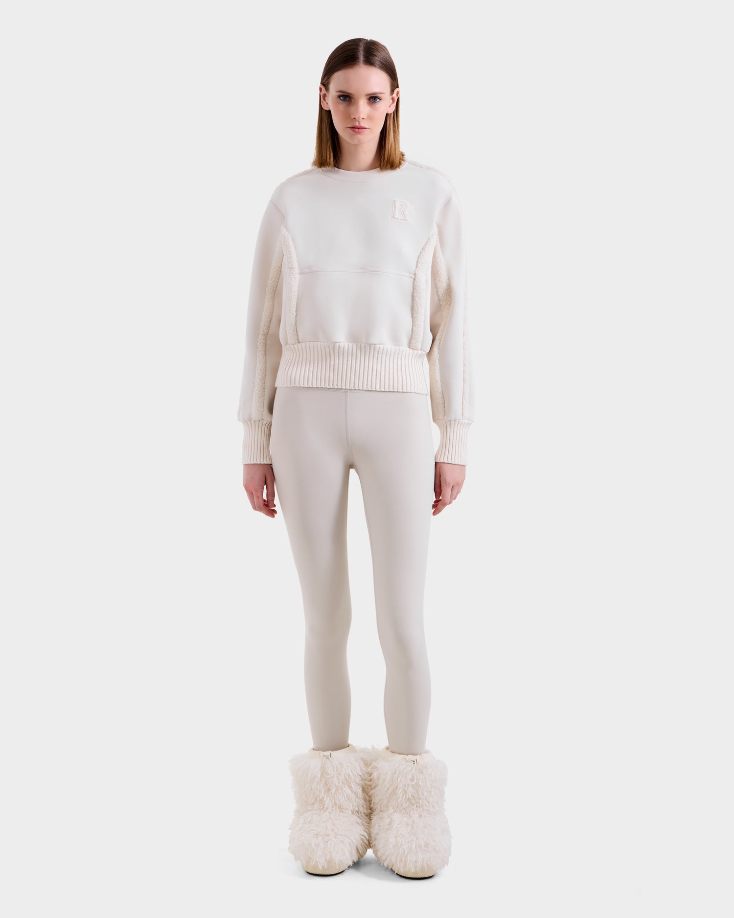 A woman stands against a white background wearing the Rudsak CHANA - 8525656 Vanilla women’s crew neck sweatshirt, matching fitted leggings, and oversized fluffy white slippers with faux shearling trim, exuding après-ski chic.