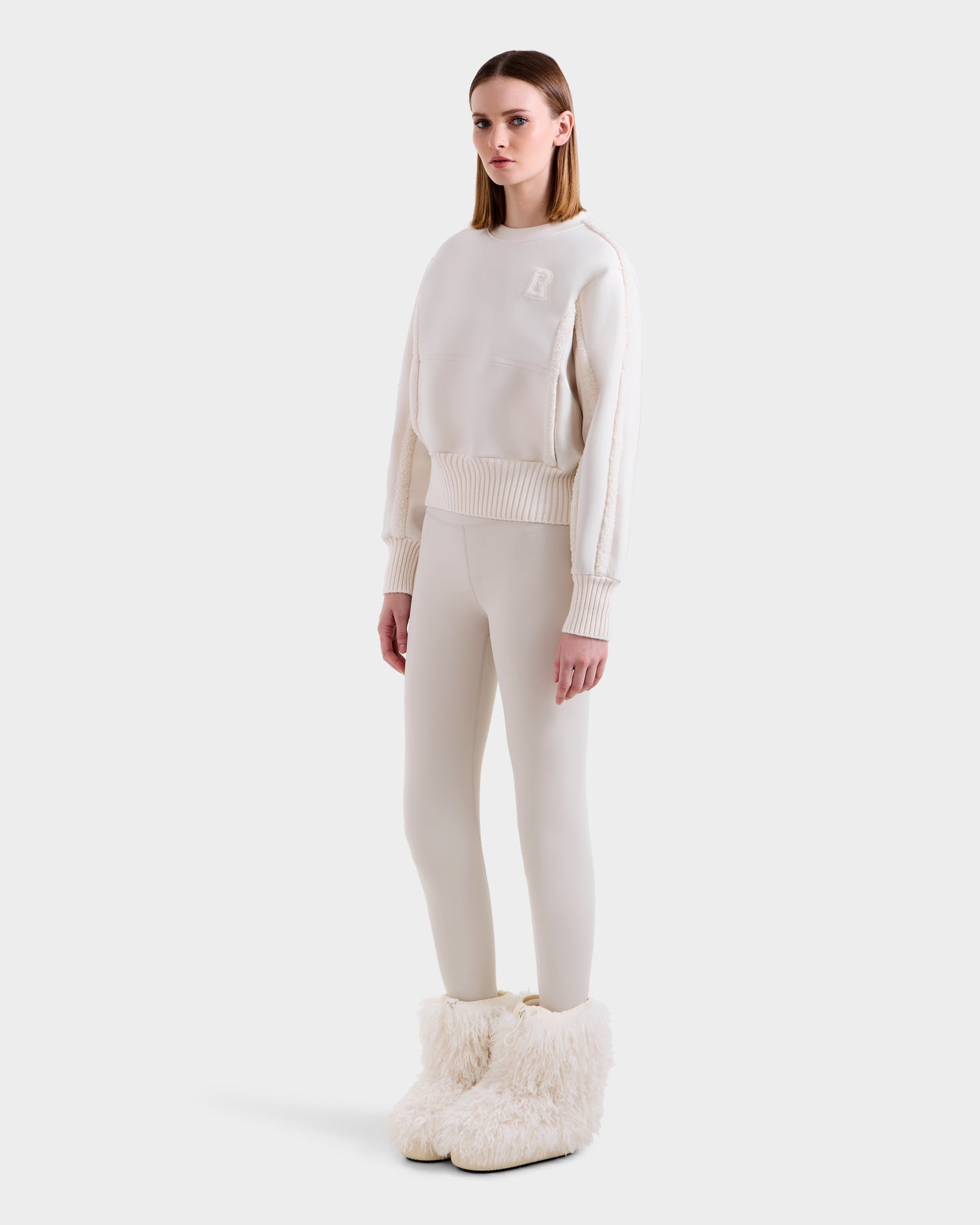 A woman with straight, shoulder-length hair and a neutral expression models the Rudsak CHANA - 8525656 Vanilla oversized fluffy white boots with faux shearling trim, styled with an off-white crew neck sweatshirt and matching leggings.