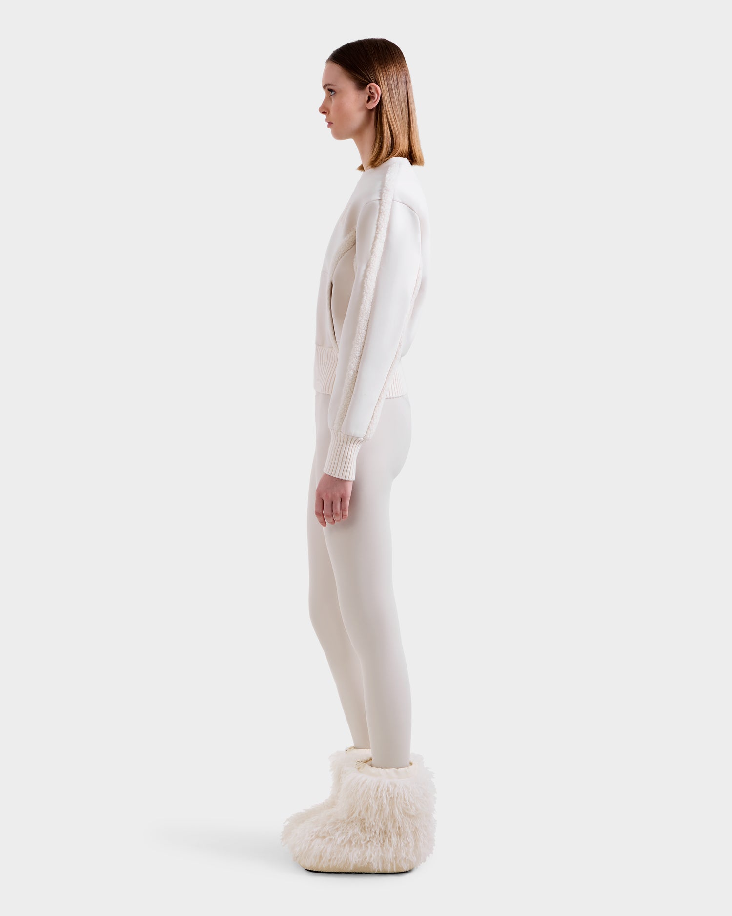 A woman in profile poses against a plain background, wearing the Rudsak CHANA - 8525656 Vanilla women's crew neck sweatshirt and matching leggings. Her straight, shoulder-length hair is parted down the middle. Fluffy slippers with faux shearling trim complete her look.