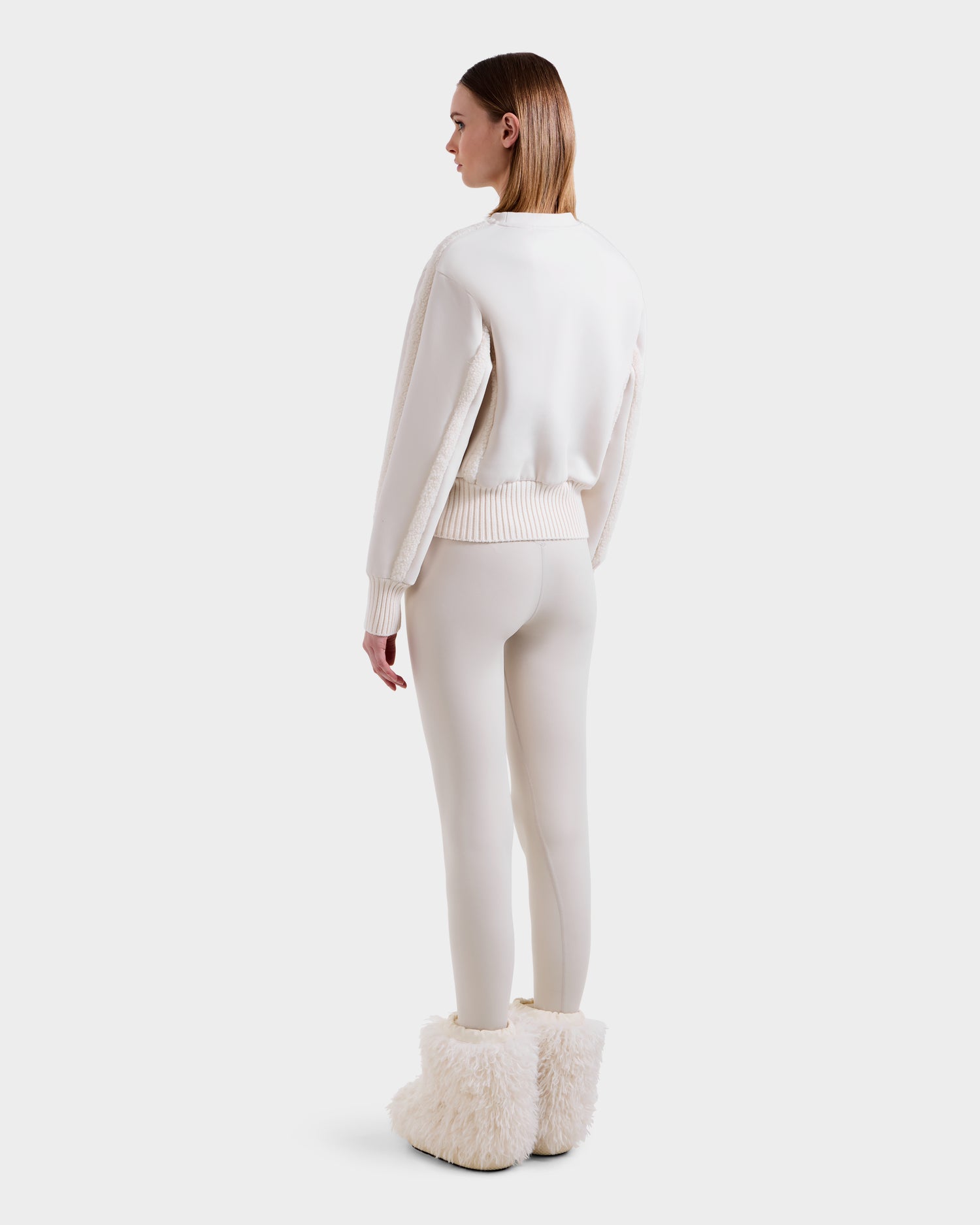 A person faces away in Rudsak’s CHANA - 8525656 Vanilla faux shearling sweatshirt, paired with leggings and fluffy white boots—effortlessly channeling après-ski chic against a plain white background.