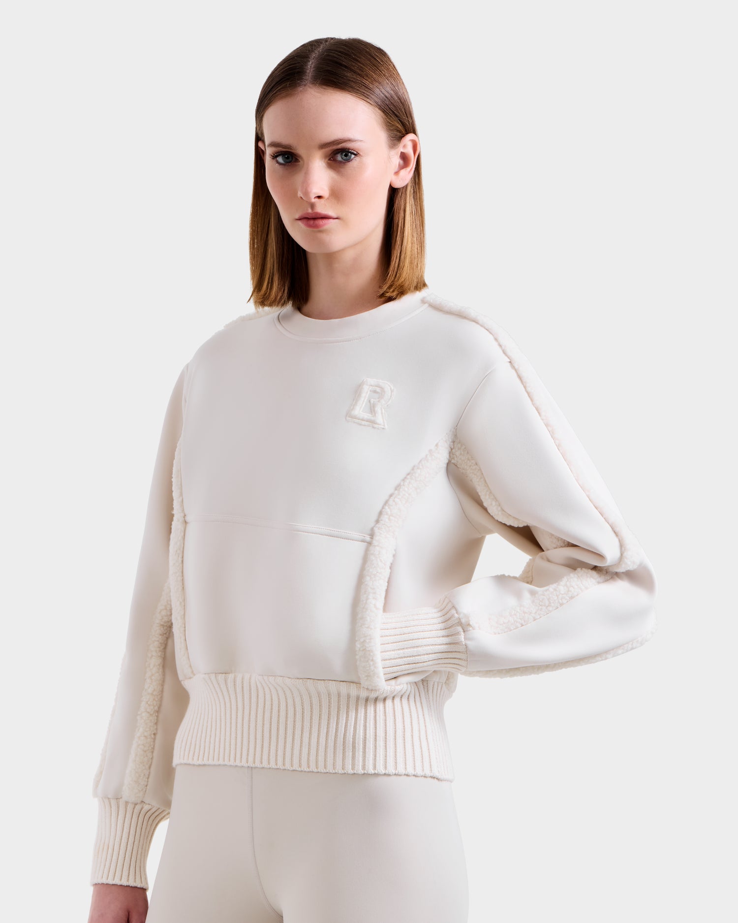 A woman with straight, shoulder-length hair wears the Rudsak CHANA - 8525656 Vanilla women’s crew neck sweatshirt featuring textured seams and an R emblem, standing against a light gray background and looking slightly to her right.