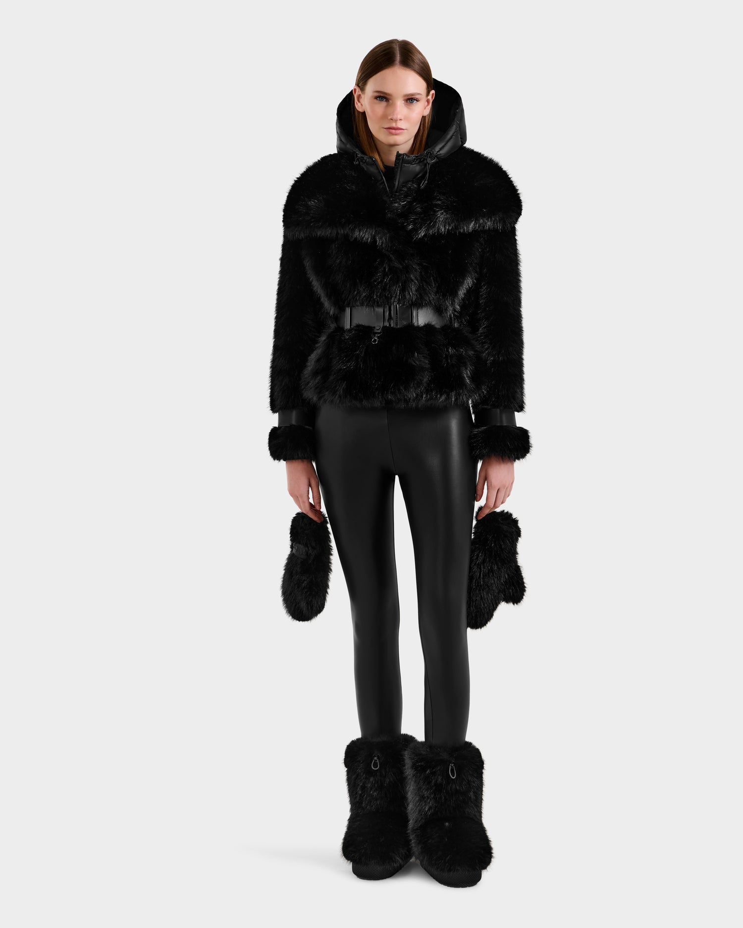 A woman faces forward in chic winter fashion, wearing the Rudsak CHLOE - 8125619 Black women's faux fur jacket with a belt, paired with black leather pants, black faux fur mittens, and boots against a plain white background.