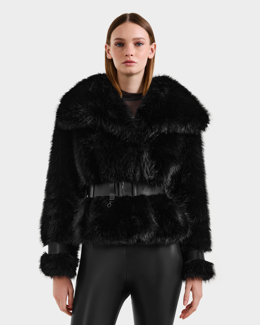 A woman with straight brown hair wears the Rudsak CHLOE - 8125619 Black faux fur jacket with a wide collar, belted waist, and leather-trimmed cuffs, paired with shiny black pants—an elegant winter wardrobe choice.
