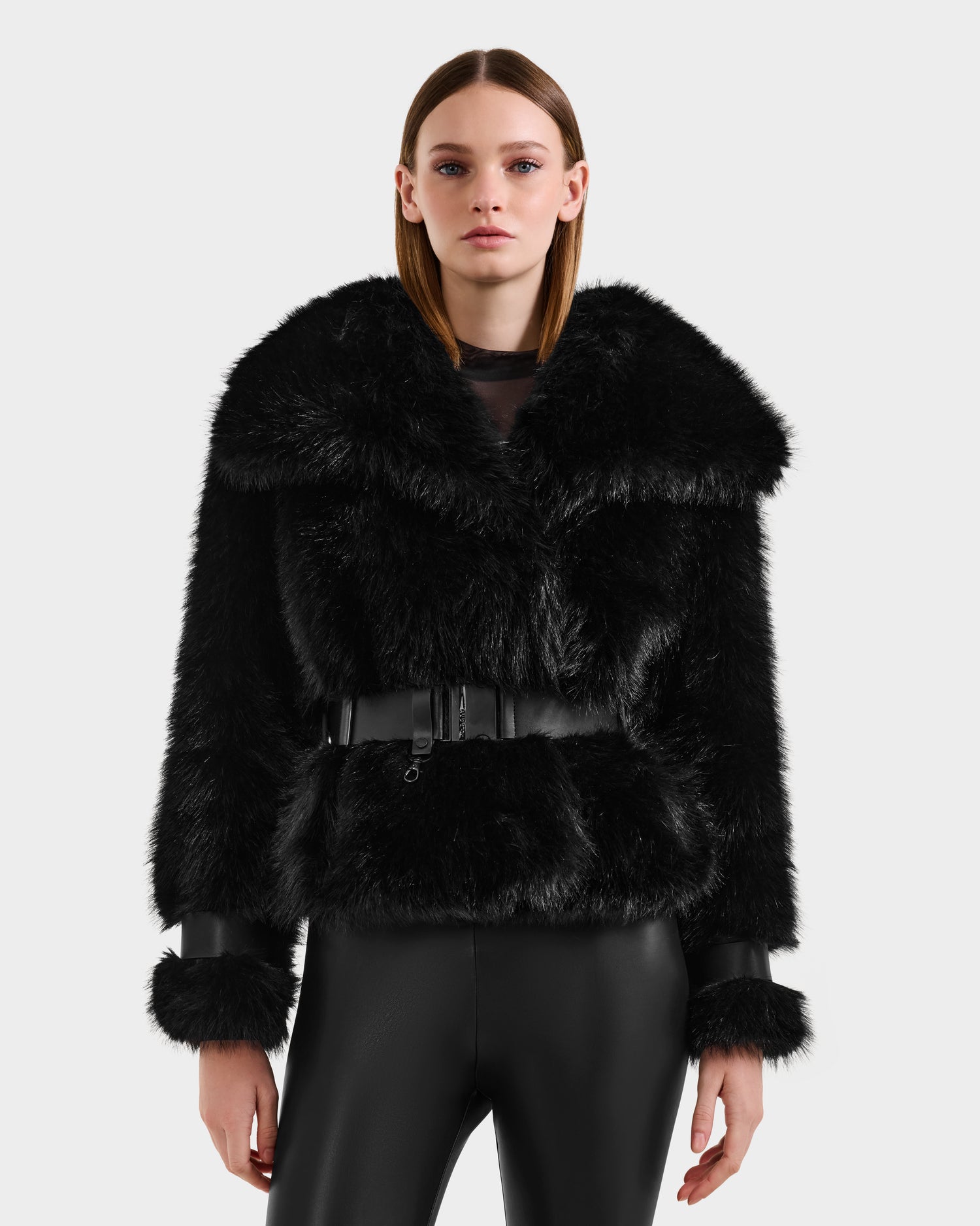 A woman with straight brown hair wears the Rudsak CHLOE - 8125619 Black faux fur jacket with a wide collar, belted waist, and leather-trimmed cuffs, paired with shiny black pants—an elegant winter wardrobe choice.