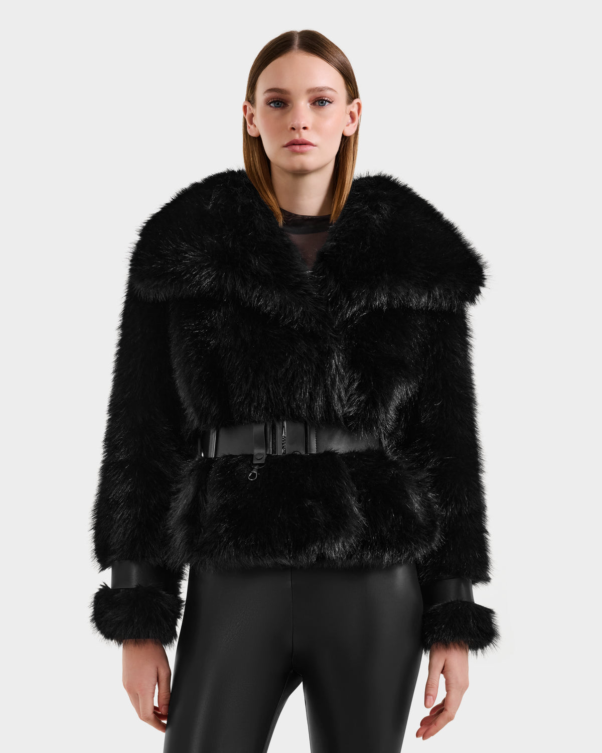 A woman with straight brown hair wears the Rudsak CHLOE - 8125619 Black faux fur jacket with a wide collar, belted waist, and leather-trimmed cuffs, paired with shiny black pants—an elegant winter wardrobe choice.