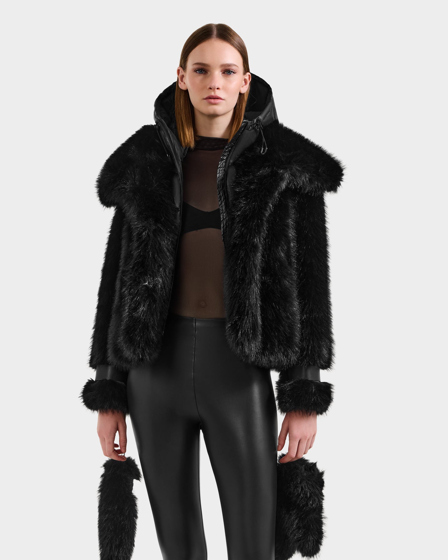 A woman with straight, shoulder-length hair and a neutral expression wears the Rudsak CHLOE - 8125619 Black women's faux fur jacket with oversized collar and cuffs over a sheer top and shiny leggings—a standout winter wardrobe piece.