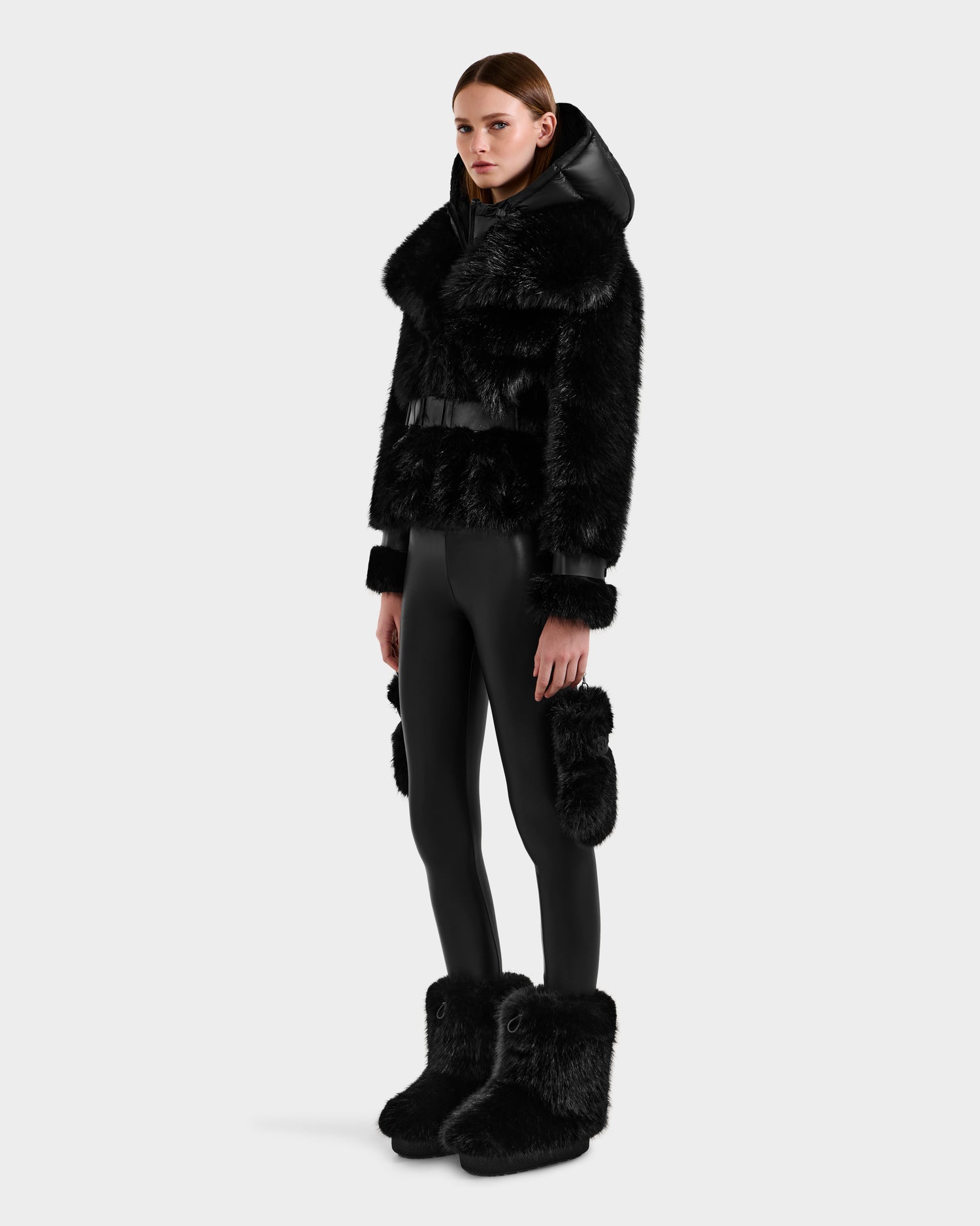 A person stands against a plain background wearing the CHLOE - 8125619 Black women's faux fur jacket by Rudsak, paired with matching fur-trimmed boots, black leggings, and fur accents—a chic winter fashion look.