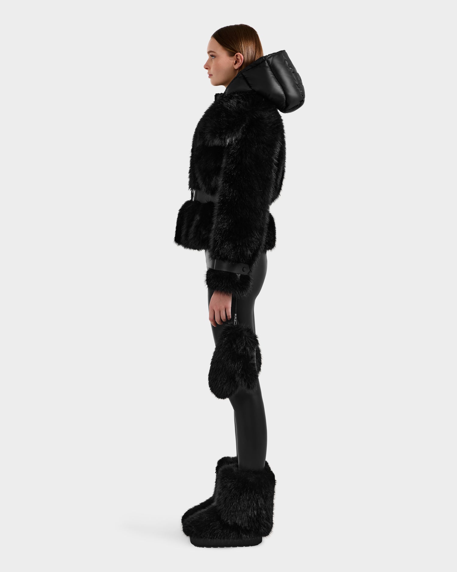 A woman stands in profile wearing the Rudsak CHLOE - 8125619 Black faux fur jacket with a hood, paired with matching faux fur boots and black leggings—an effortlessly chic winter look against a plain light background.