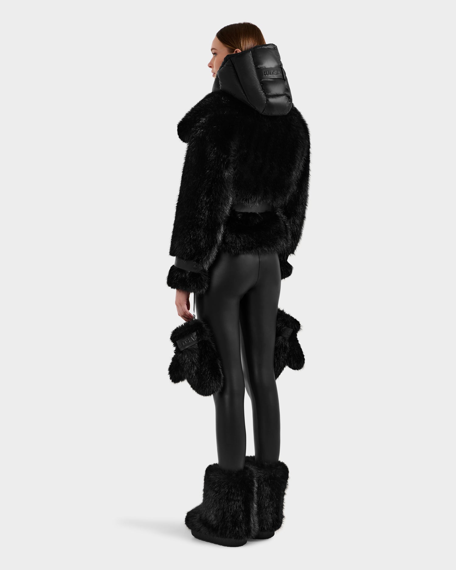 A woman faces away, modeling Rudsak's CHLOE - 8125619 Black faux fur jacket with large collar and hood, black vegan leather leggings, matching mittens, and black faux fur boots against a light gray background.
