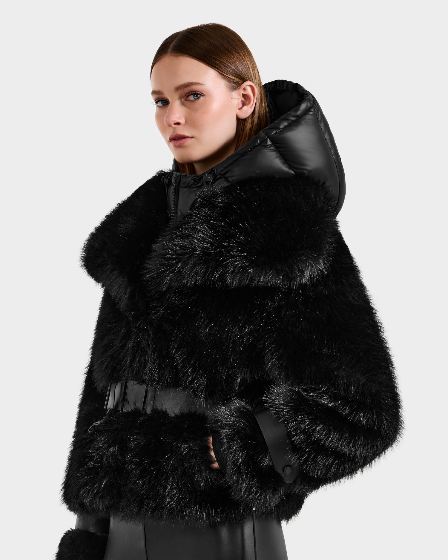 A woman in the Rudsak CHLOE - 8125619 Black faux fur jacket with a large hood and vegan leather accents stands against a light background, looking to the side—a chic winter fashion statement.