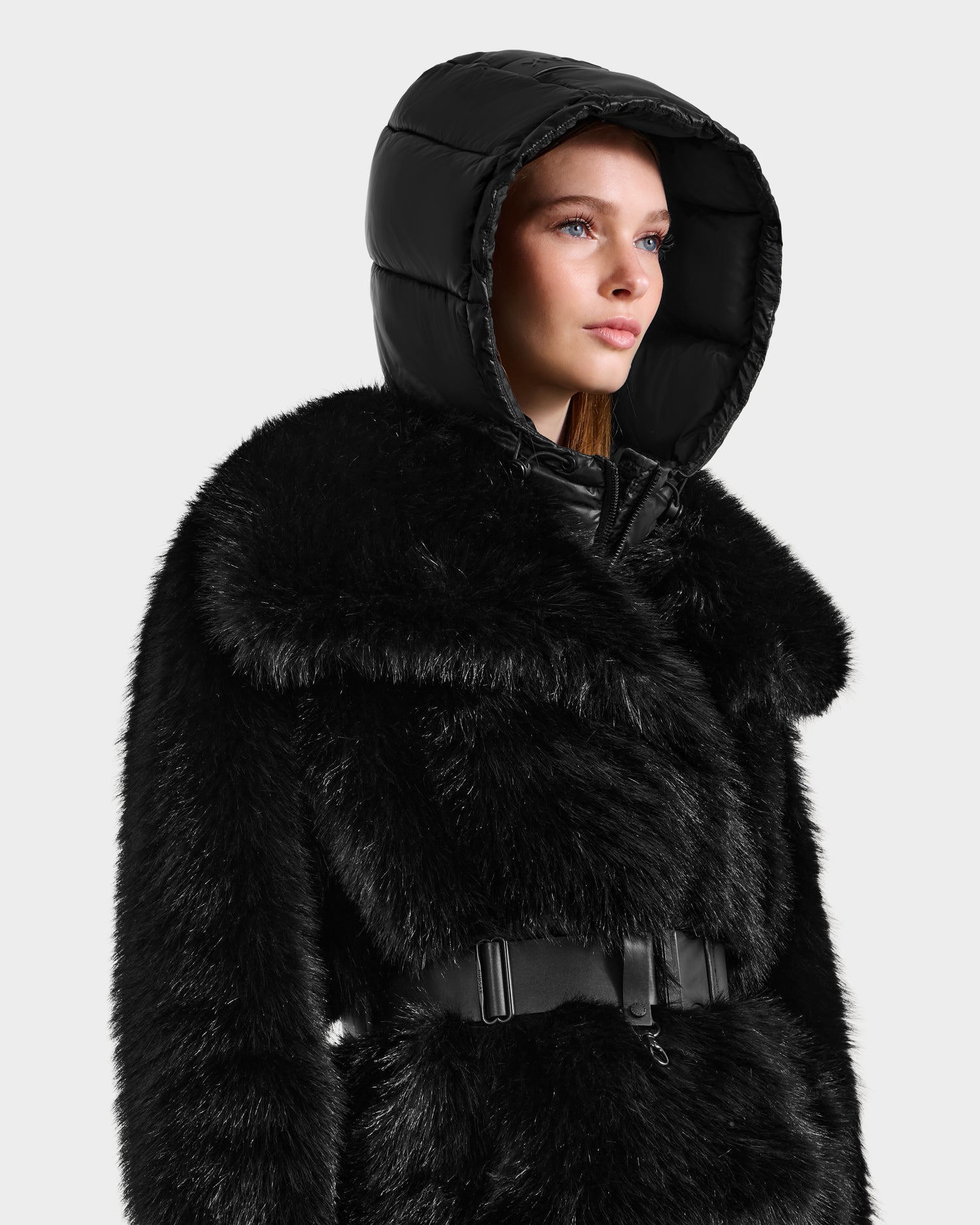 A woman in the CHLOE - 8125619 Black women's faux fur jacket by Rudsak, featuring a matching belt and shiny hood, looks confidently to the side against a white background—ideal for chic winter style.