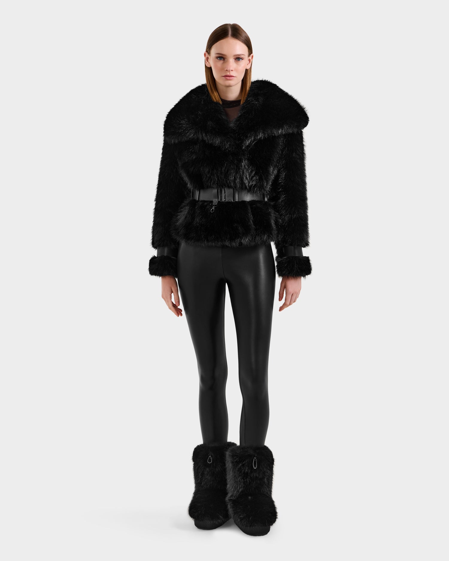 A model faces forward wearing the Rudsak CHLOE - 8125619 Black women's faux fur jacket with a wide collar, black belted waist, matching faux fur boots, and shiny black leggings—an ultra-chic winter look against a light gray background.