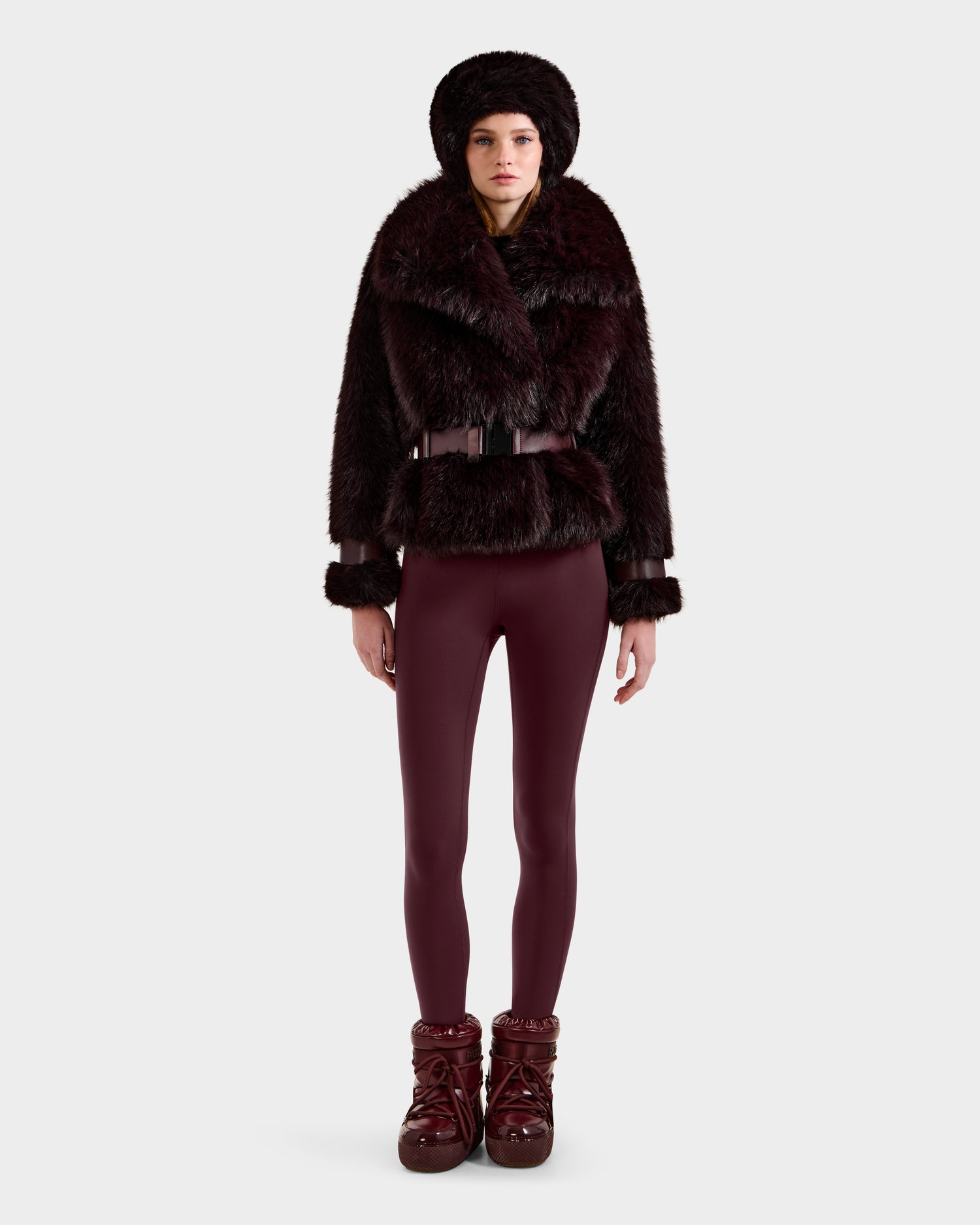 A person stands facing forward in a Rudsak CHLOE - 8125619 Bordeaux faux fur jacket with a wide collar, matching fur hat, fitted burgundy leggings, and snow boots. The background is plain light gray.