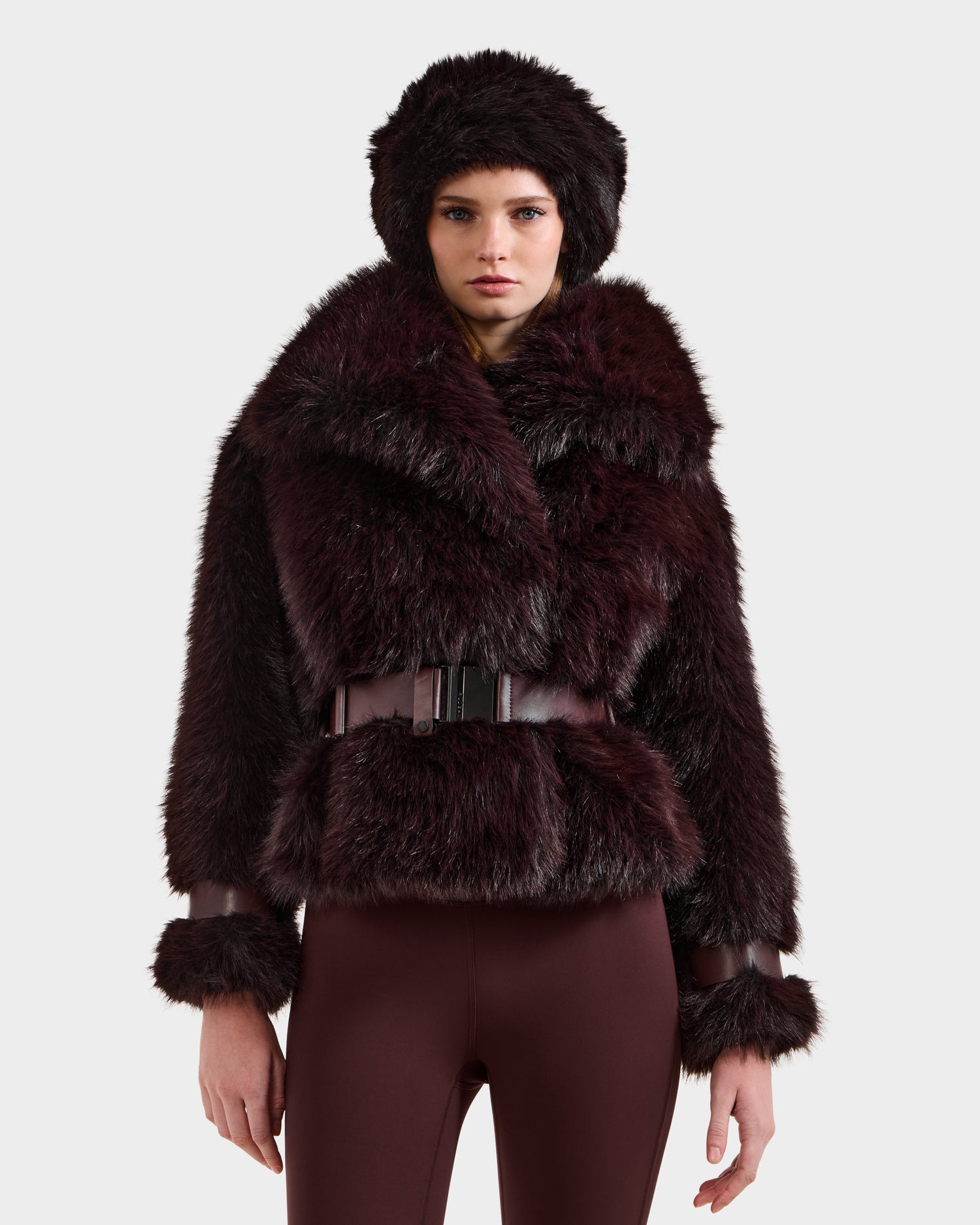 A woman models the Rudsak CHLOE - 8125619 Bordeaux: a dark brown faux fur jacket with a wide collar, matching hat, and fitted maroon pants—perfect for elevating any winter wardrobe.