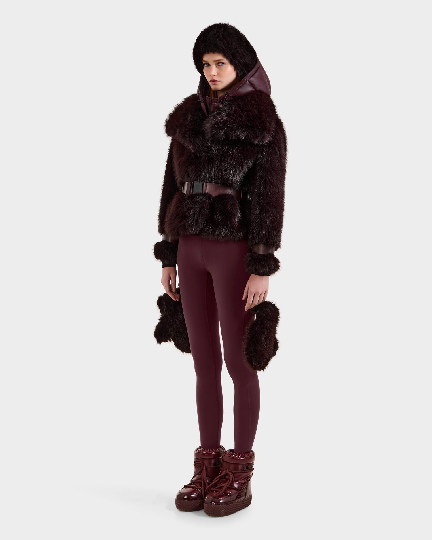 A person stands against a plain white background, wearing the Rudsak CHLOE - 8125619 Bordeaux women's faux fur jacket with a belt, matching leggings, and chunky burgundy boots. Plush fur accents on the cuffs and hips complete the stylish look.