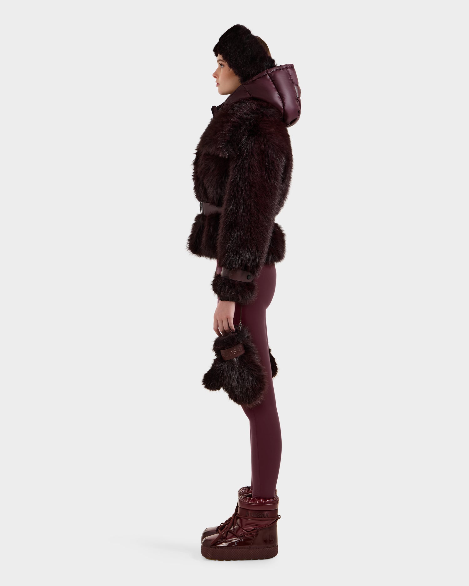 A person in side profile models the CHLOE - 8125619 Bordeaux faux fur jacket by Rudsak with matching pants, fur-trimmed gloves, a hat, and boots—an elegant winter look—against a plain white background.