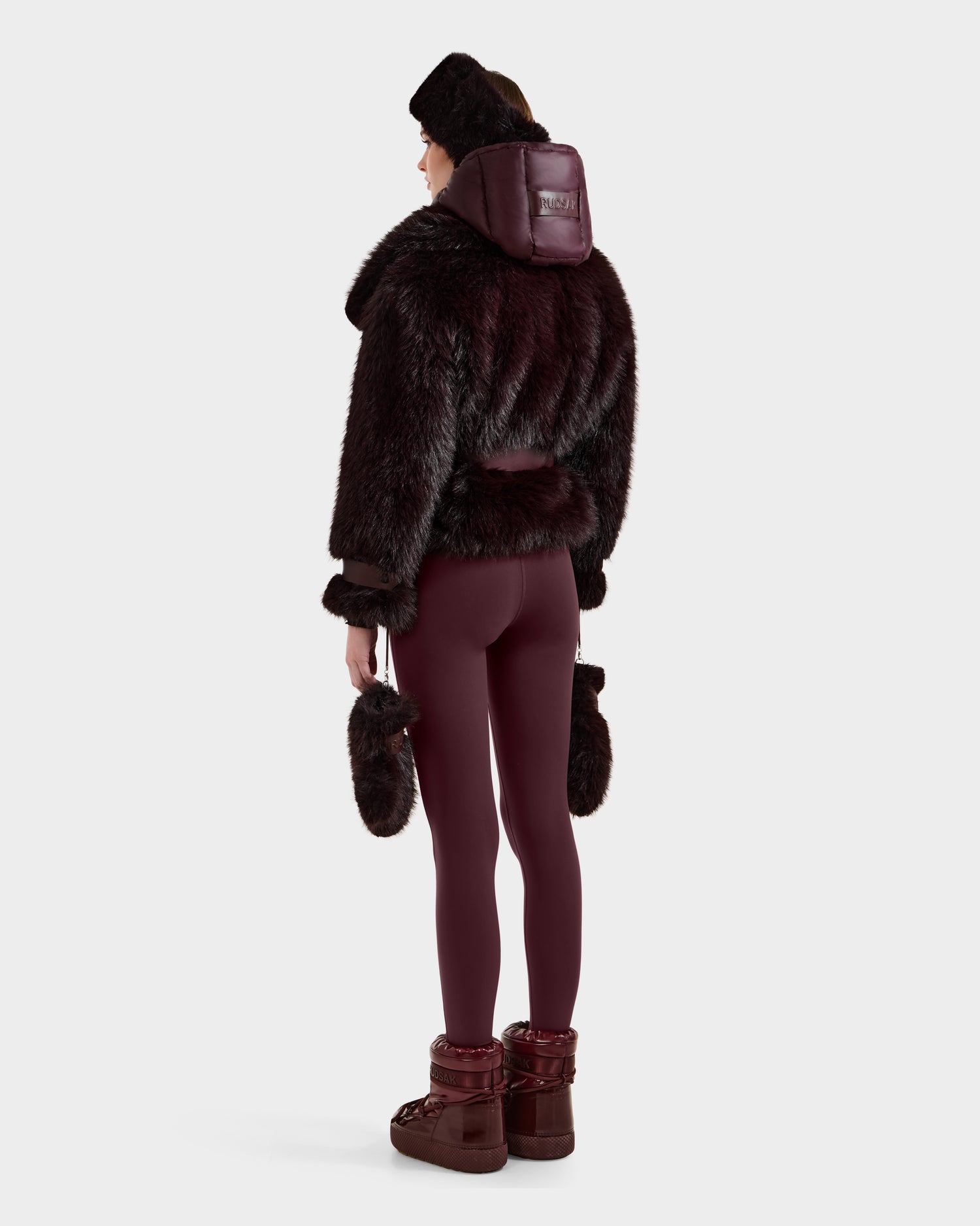 A person models the Rudsak CHLOE - 8125619 Bordeaux faux fur jacket with matching hat, leggings, and fur-trimmed boots, standing against a white background to highlight a stylish winter look.