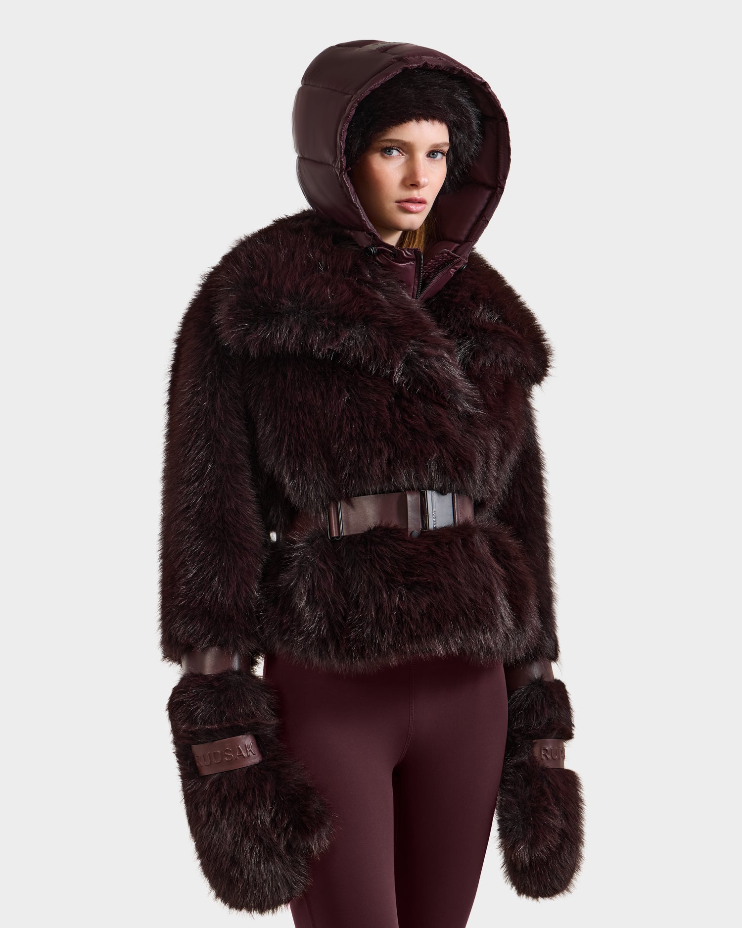 A woman in the Rudsak CHLOE - 8125619 Bordeaux dark brown faux fur jacket with a shiny hood, furry mittens, and burgundy fitted pants poses against a plain background, highlighting a chic winter look.