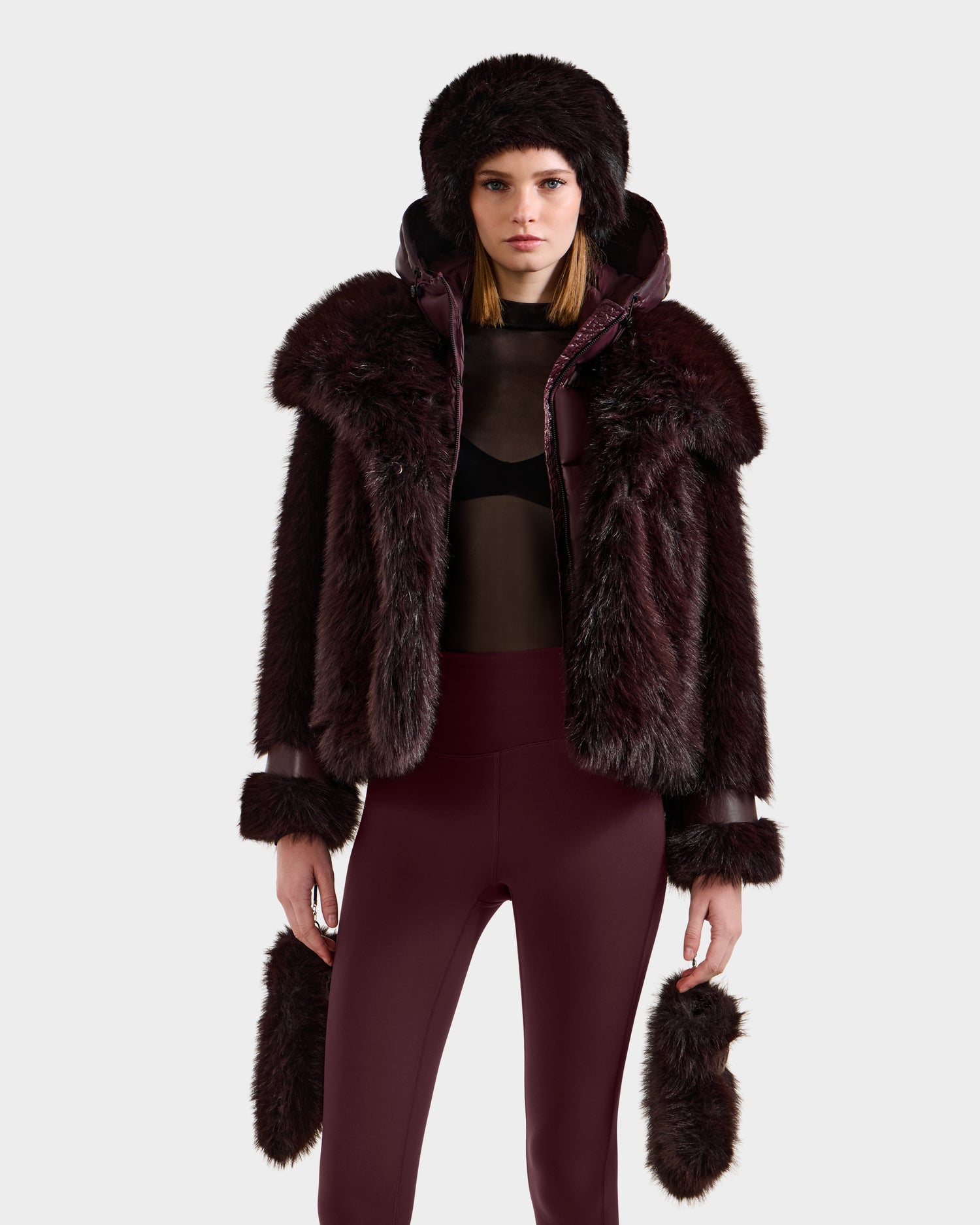 A woman showcases the Rudsak CHLOE - 8125619 Bordeaux faux fur jacket with a matching fur-trimmed hood, cuffs, and mittens, paired with high-waisted burgundy leggings and a sheer black top against a plain light background.