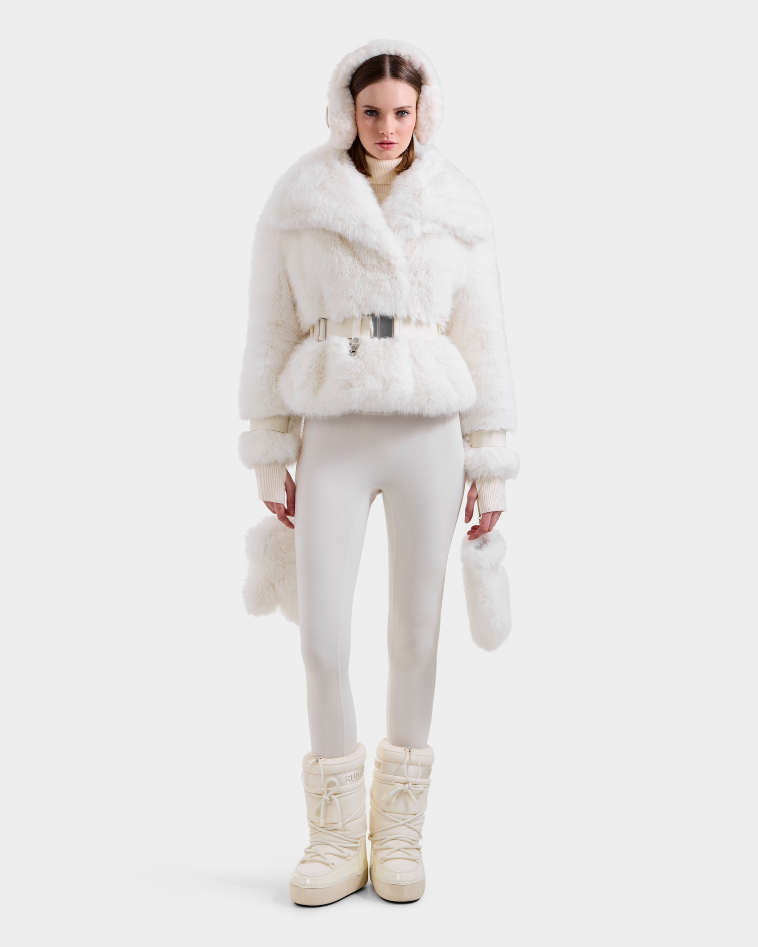 A woman models the Rudsak CHLOE - 8125619 Iceland in chic winter style—an all-white ensemble with a fluffy women's faux fur jacket and hood, matching mittens, fitted pants, and puffy snow boots against a plain white backdrop.
