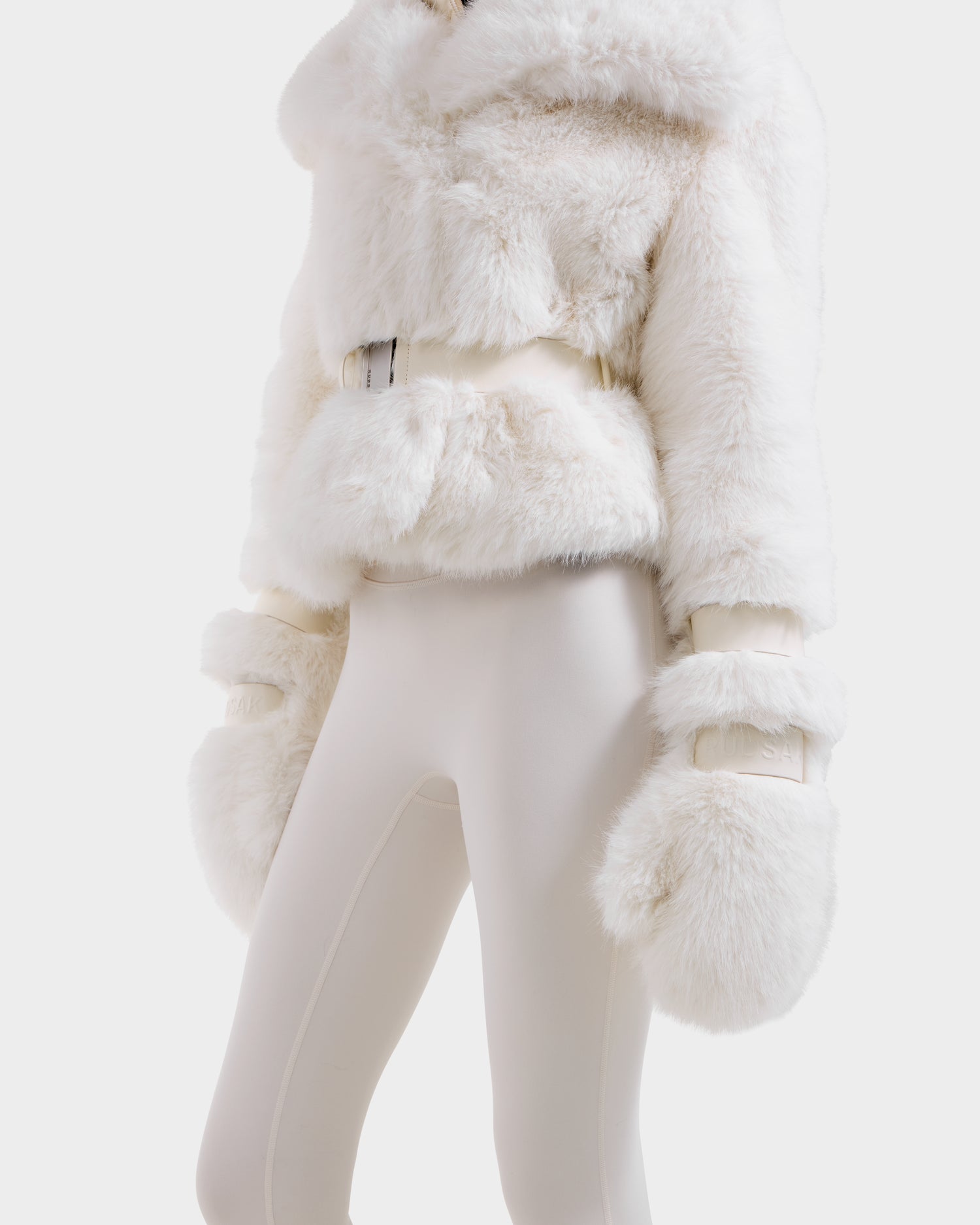 A person models the Rudsak CHLOE - 8125619 Iceland women's off-white faux fur jacket with a matching belt and plush sleeves, styled over white fitted pants. The winter-ready look is set against a plain white background, face not shown.