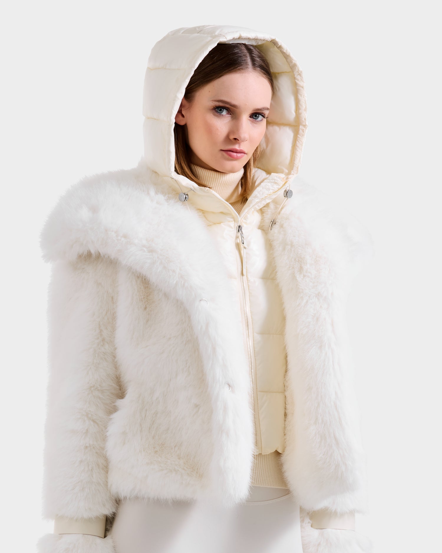 A woman models winter fashion in the Rudsak CHLOE - 8125619 Iceland faux fur jacket over a quilted white hooded jacket, hood up, standing against a plain light background with her light brown hair framing her neutral expression.