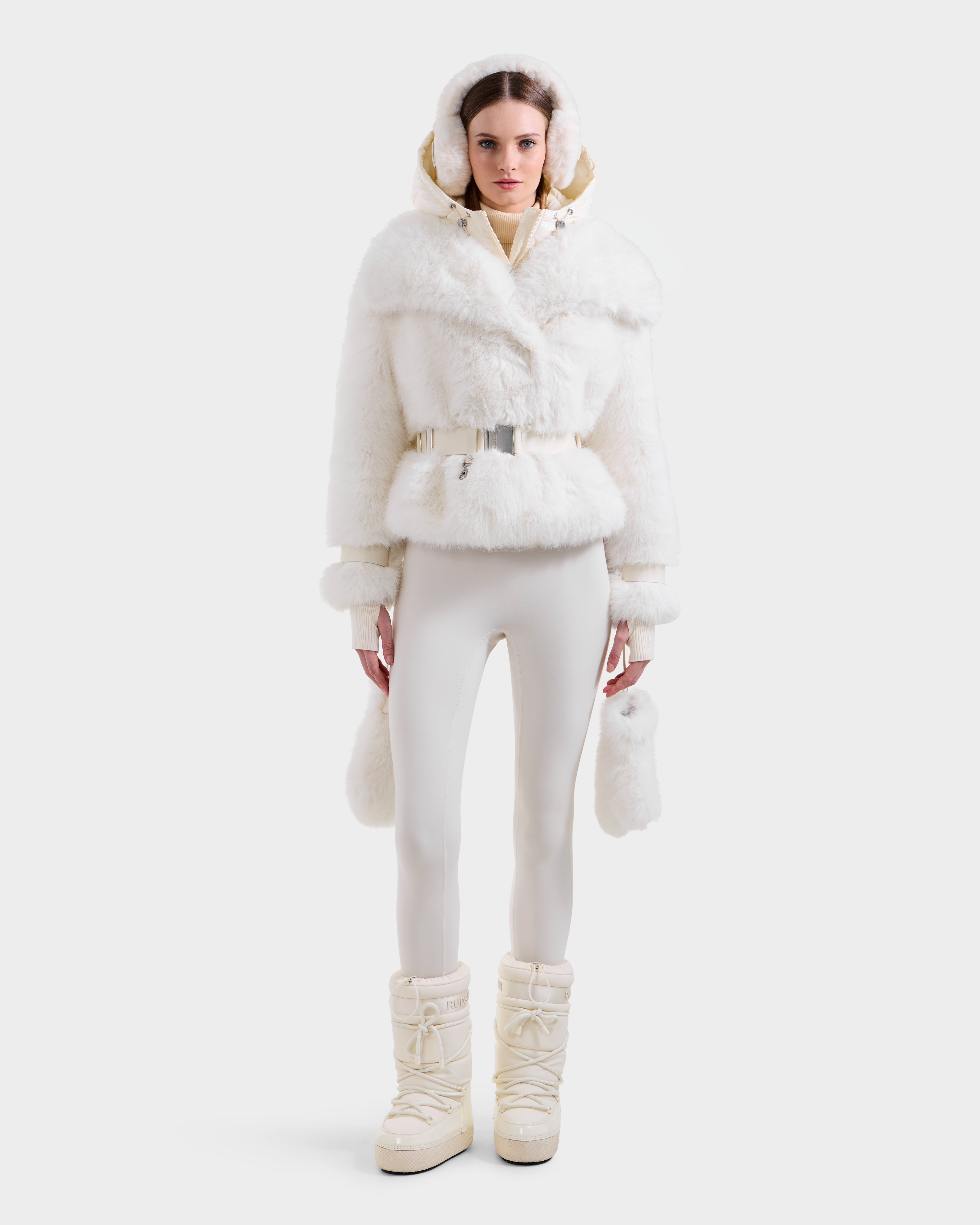 A woman faces forward in the Rudsak CHLOE - 8125619 Iceland faux fur jacket with hood, paired with white fitted pants, winter boots, and a small furry purse—ideal for chic winter style against a plain background.