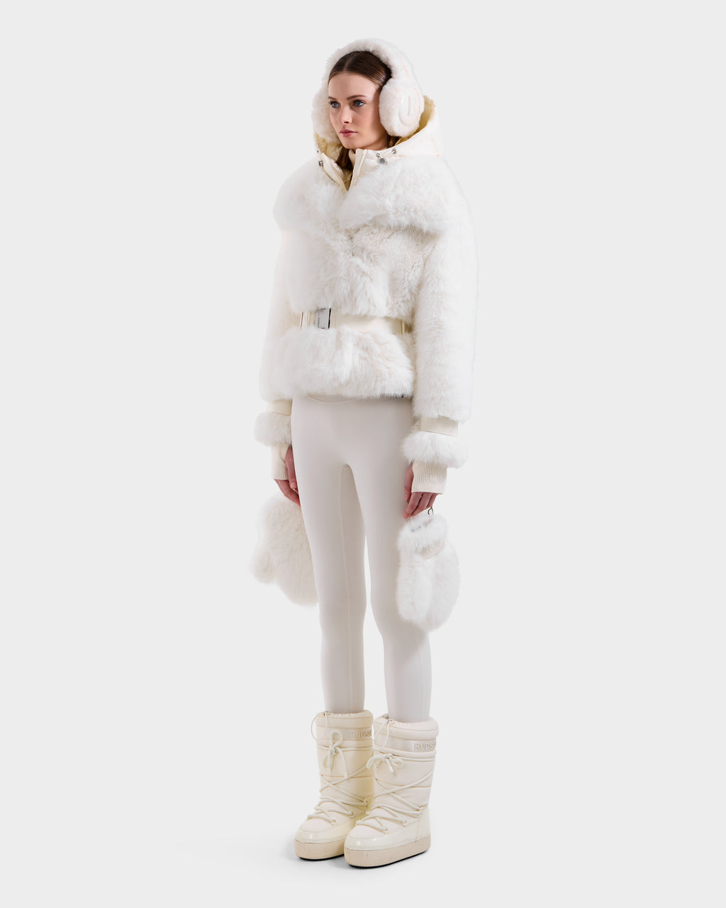 A woman models the Rudsak CHLOE - 8125619 Iceland white faux fur hooded jacket with matching earmuffs, fitted white pants, and cream snow boots for an effortlessly chic winter look against a plain white background.