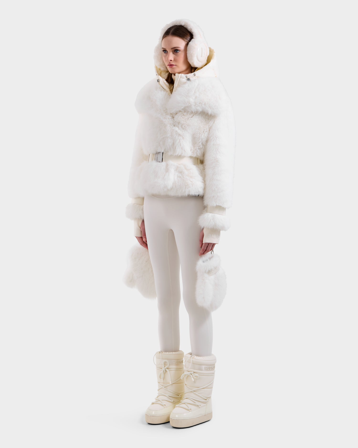 A woman models the Rudsak CHLOE - 8125619 Iceland white faux fur hooded jacket with matching earmuffs, fitted white pants, and cream snow boots for an effortlessly chic winter look against a plain white background.