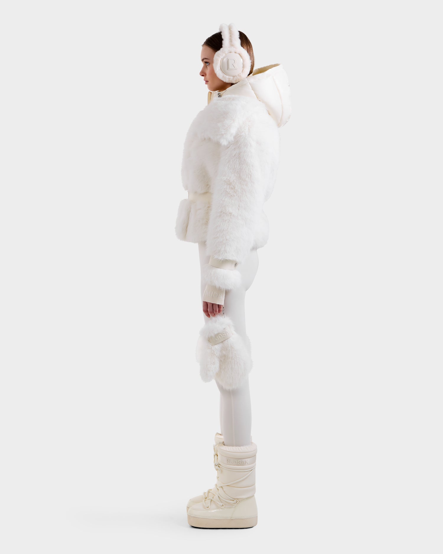 A woman stands in profile wearing the Rudsak CHLOE - 8125619 Iceland faux fur jacket with a fluffy white hood, paired with white fitted pants, furry mittens, and cream winter boots—showcasing chic style against a light background.