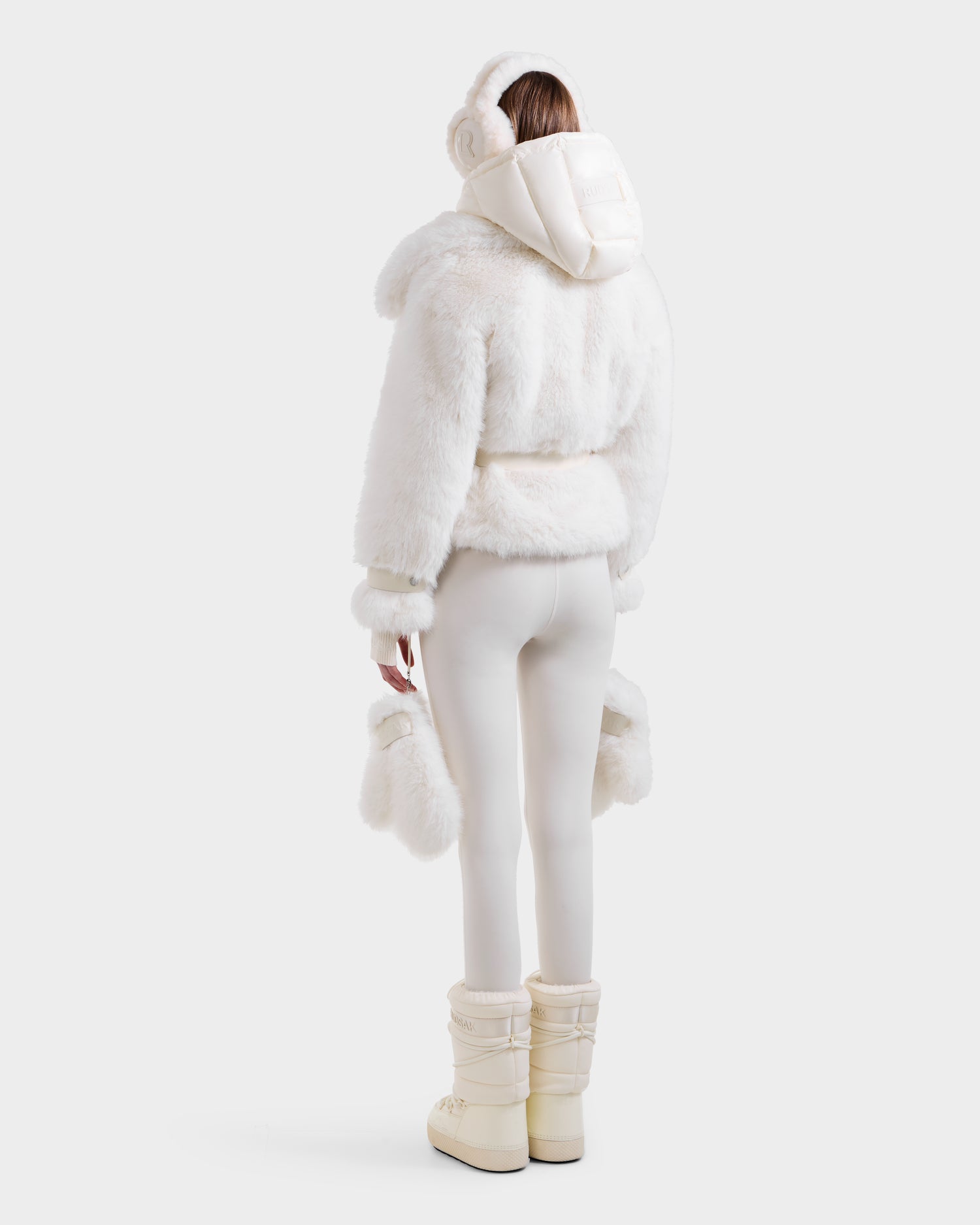 A woman models the Rudsak CHLOE - 8125619 Iceland women’s faux fur jacket with furry hood, white earmuffs, fitted white pants, fur-trimmed mittens, and ivory snow boots—showcasing chic winter style against a plain background.