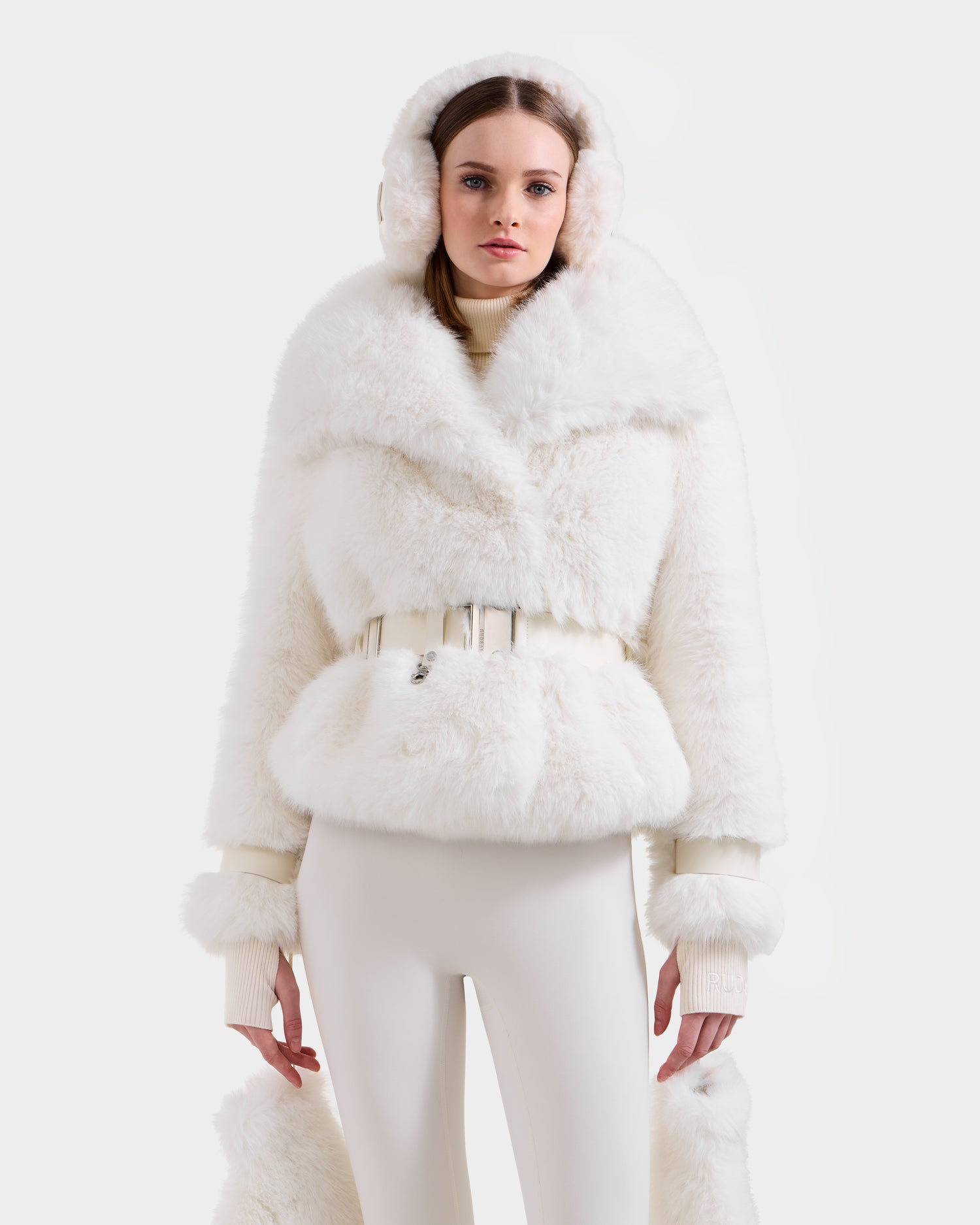 A woman stands against a plain white background wearing the Rudsak CHLOE - 8125619 Iceland: an all-white winter outfit with a women's faux fur jacket featuring a fluffy hood, wide collar, matching ear muffs, and high-waisted pants.