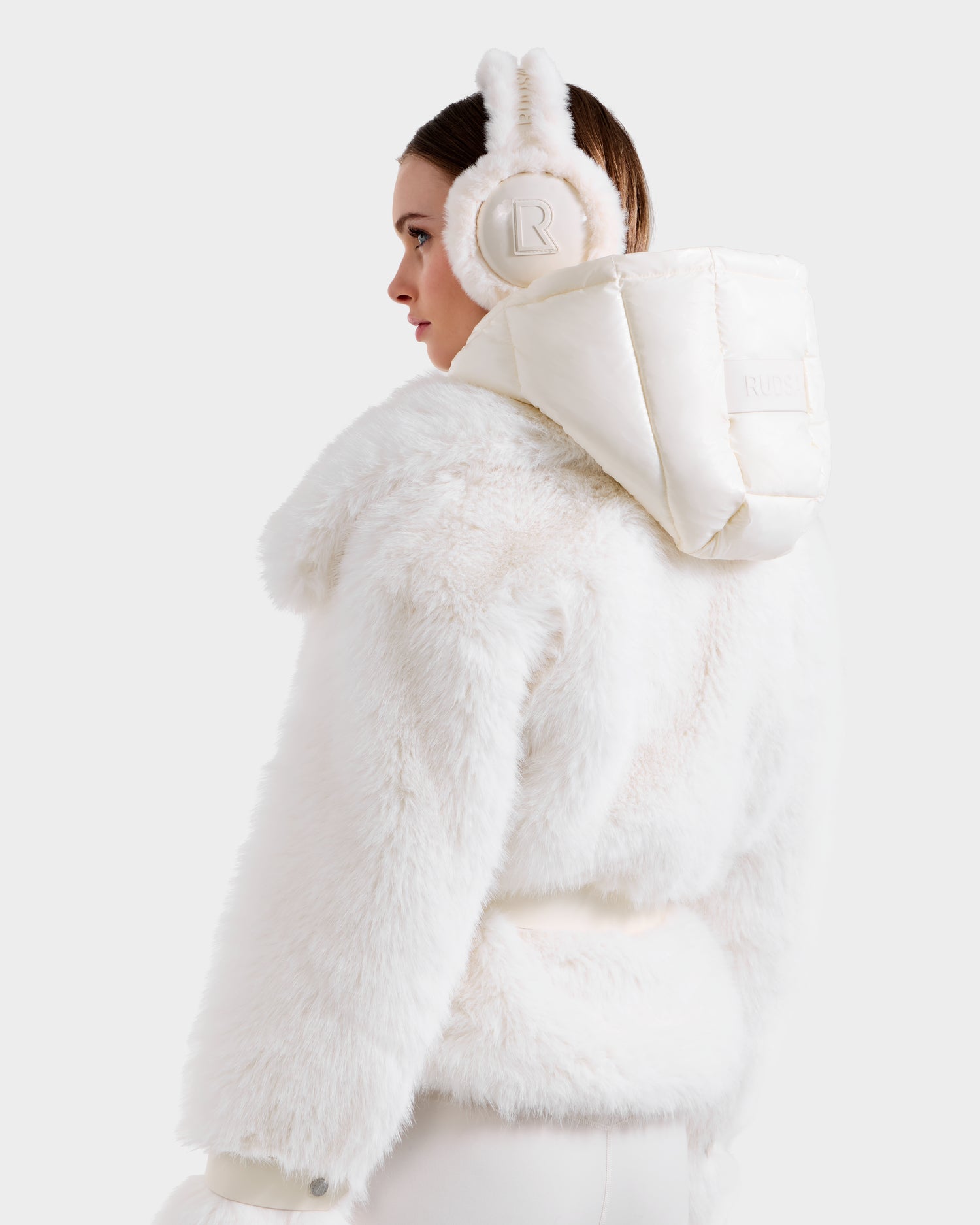 A woman models the Rudsak CHLOE - 8125619 Iceland faux fur jacket, paired with fluffy bunny-ear earmuffs and a padded hood, shown in profile against a simple light background.