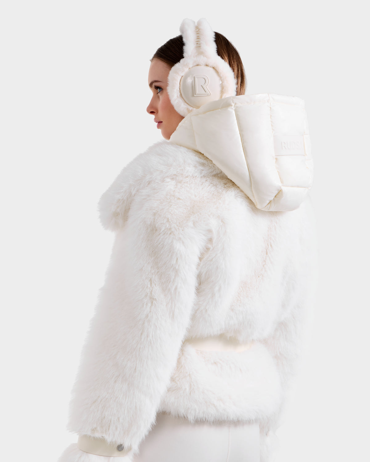 A woman models the Rudsak CHLOE - 8125619 Iceland faux fur jacket, paired with fluffy bunny-ear earmuffs and a padded hood, shown in profile against a simple light background.