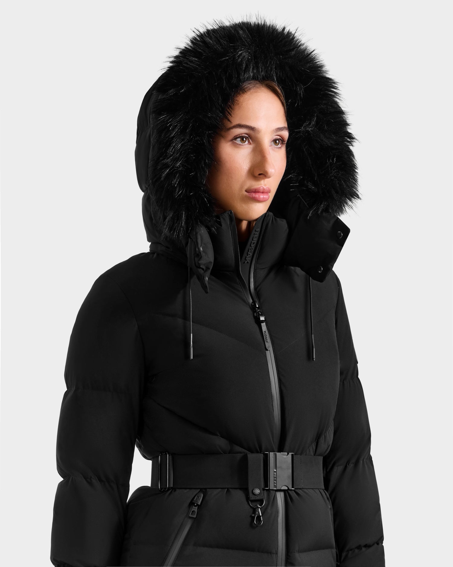 A person models the Rudsak CHRISTELLE M - L125542 Black jacket, a belted, weather-resistant coat with a fur-lined hood, shown against a plain light background.